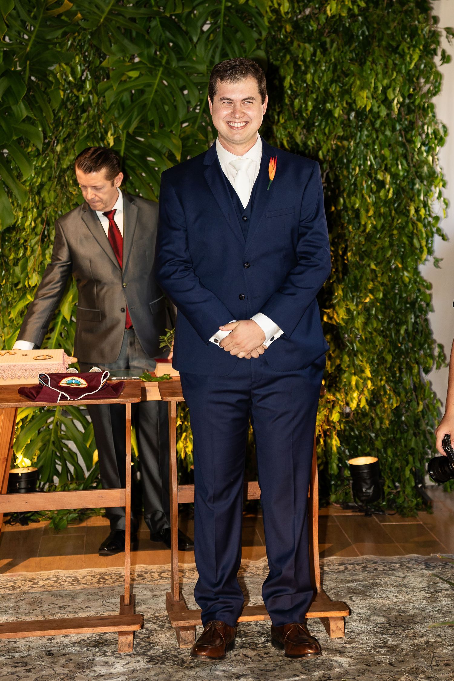 Man in navy blue suit with orange boutonniere smiling at formal event with green plant wall backdrop.