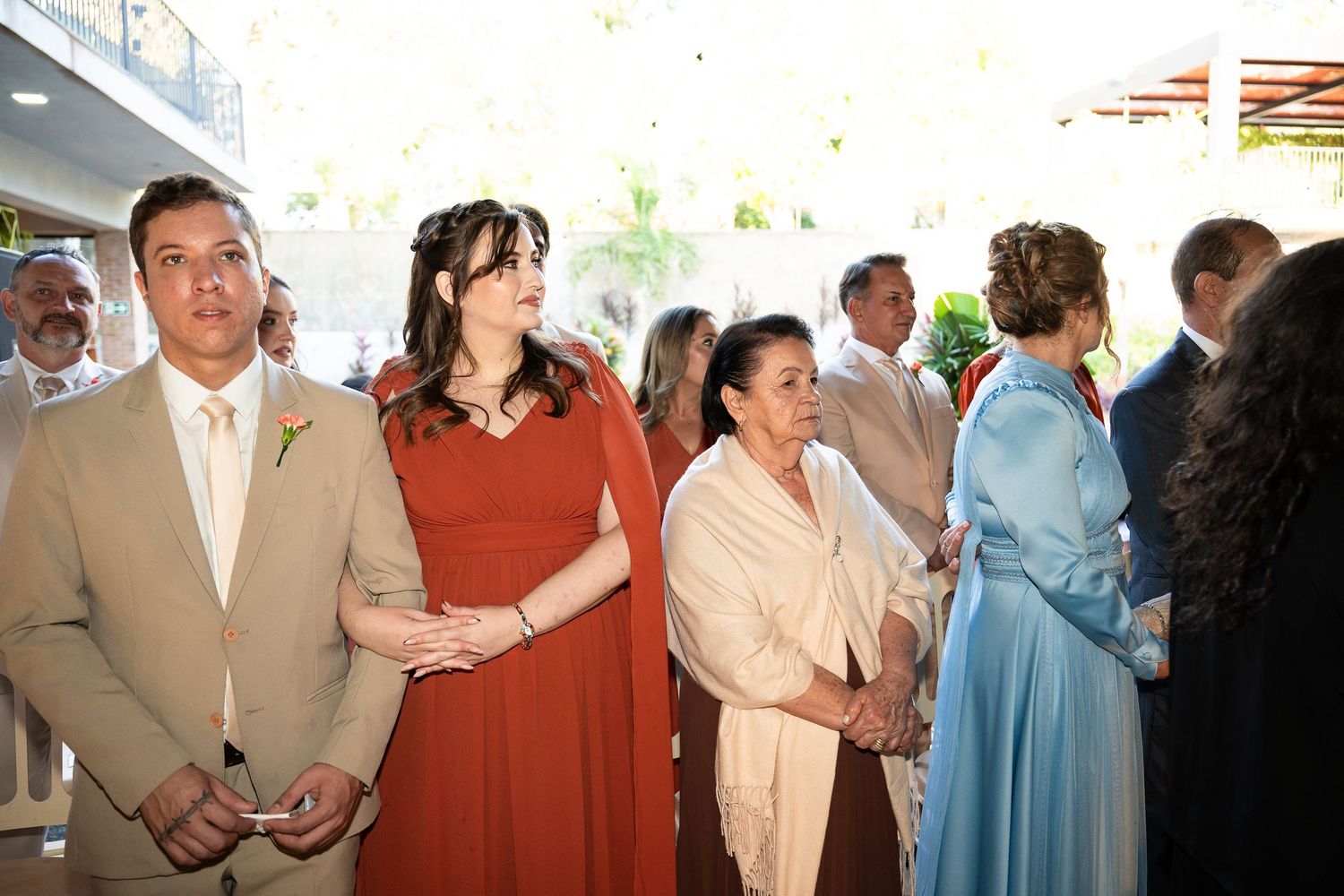 Wedding guests in formal attire stand together, including a man in tan suit and woman in rust dress, at an elegant outdoor venue.