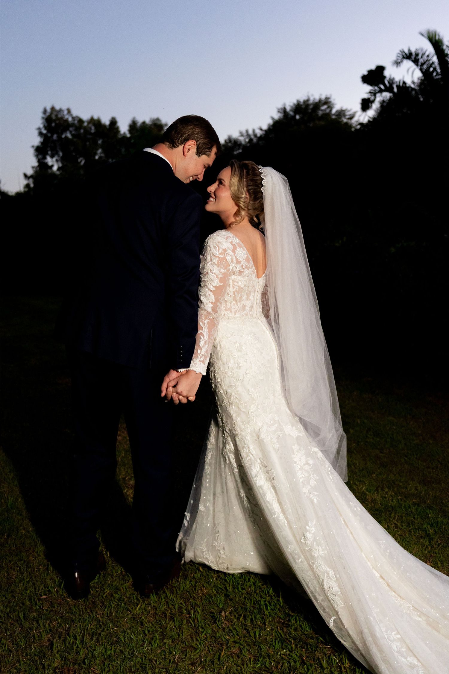 Bride in lace gown and veil holds hands with groom in navy suit at twilight outdoor wedding.