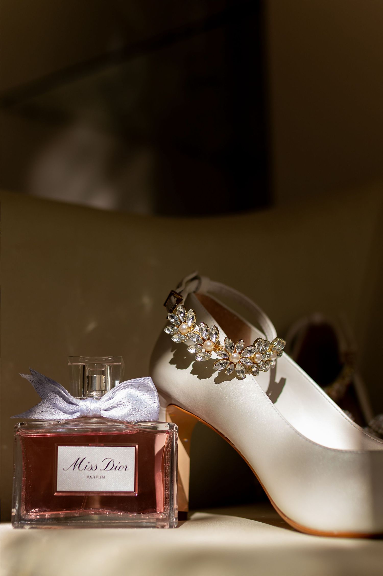 Miss Dior perfume bottle beside elegant white satin bridal heels adorned with crystal floral embellishments on a golden surface.