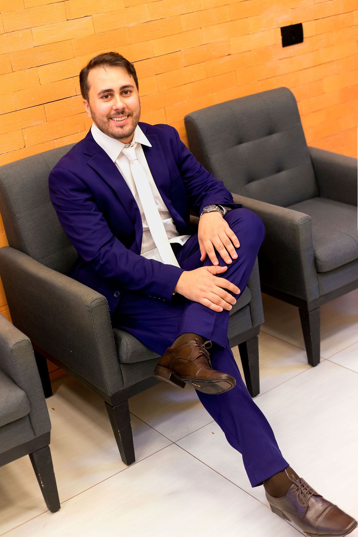Man in blue suit sitting relaxed in gray armchair, smiling confidently in modern lobby setting.