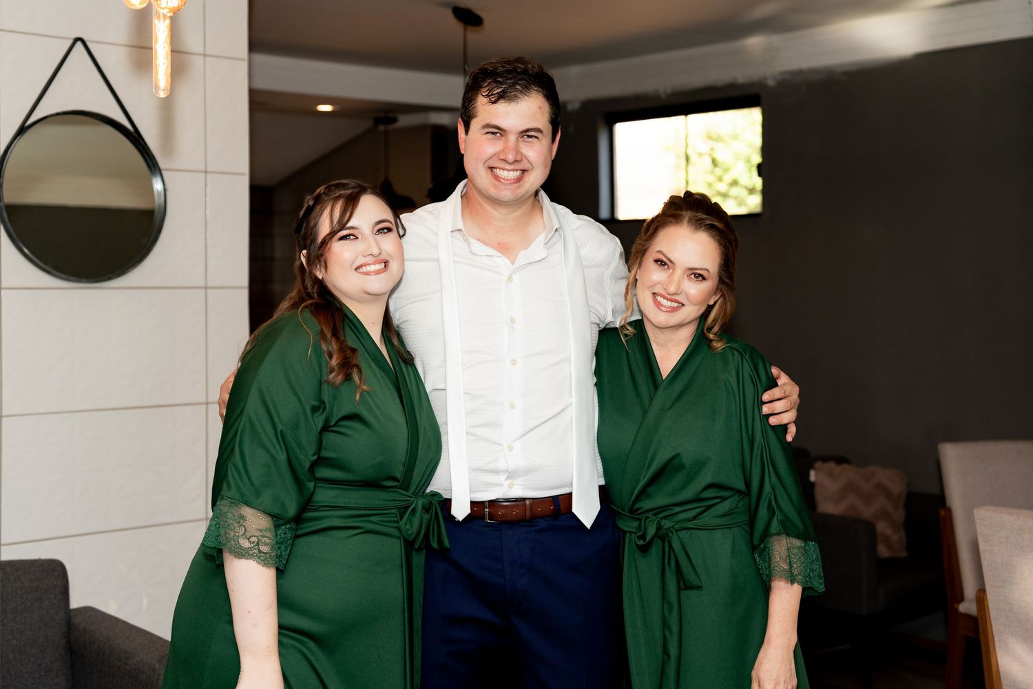 Man in white shirt posing with two women in emerald green satin robes, smiling indoors at a wedding preparation.