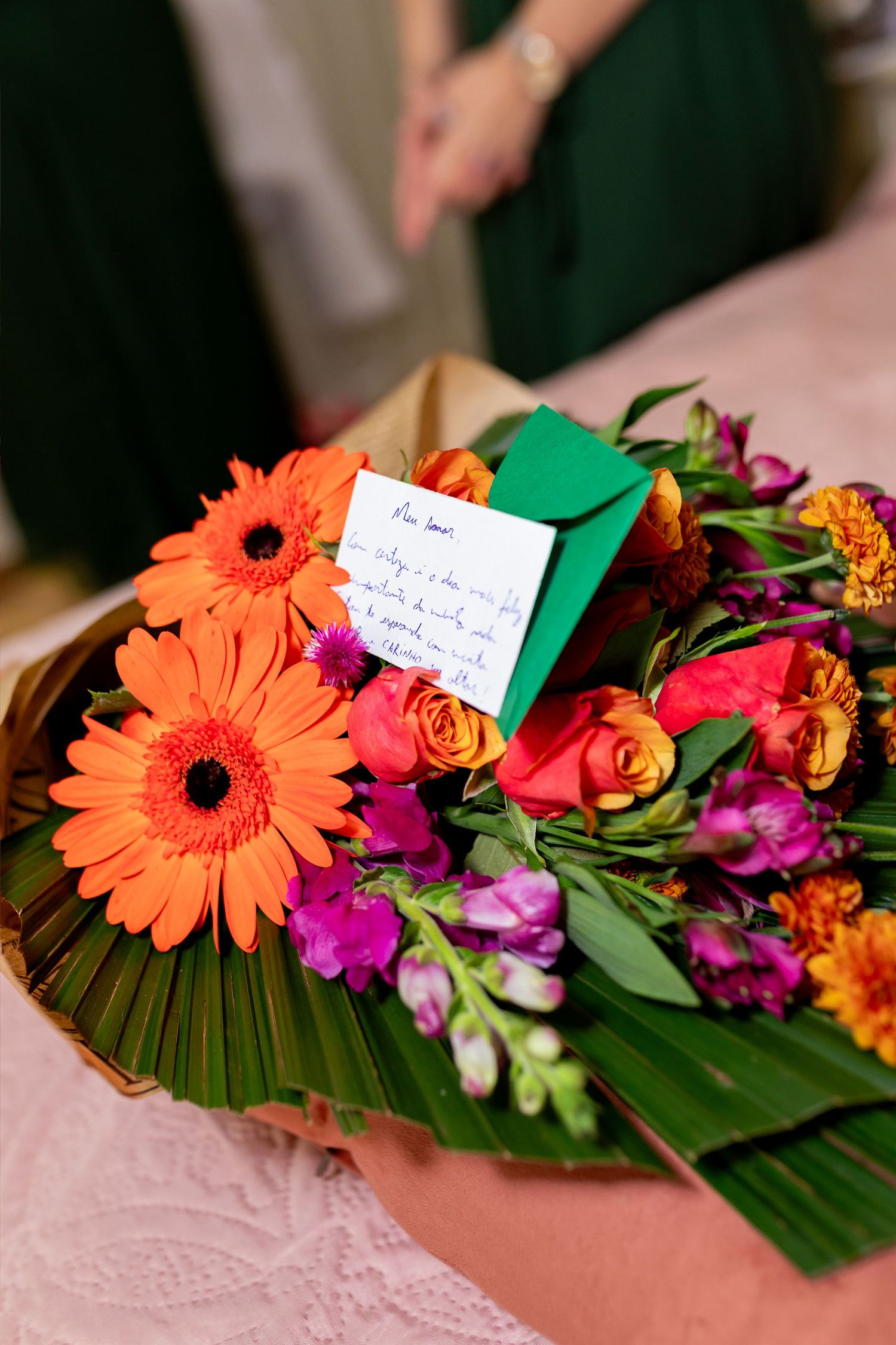 Vibrant bouquet of orange gerberas, pink roses, and purple flowers with a handwritten note and green envelope.