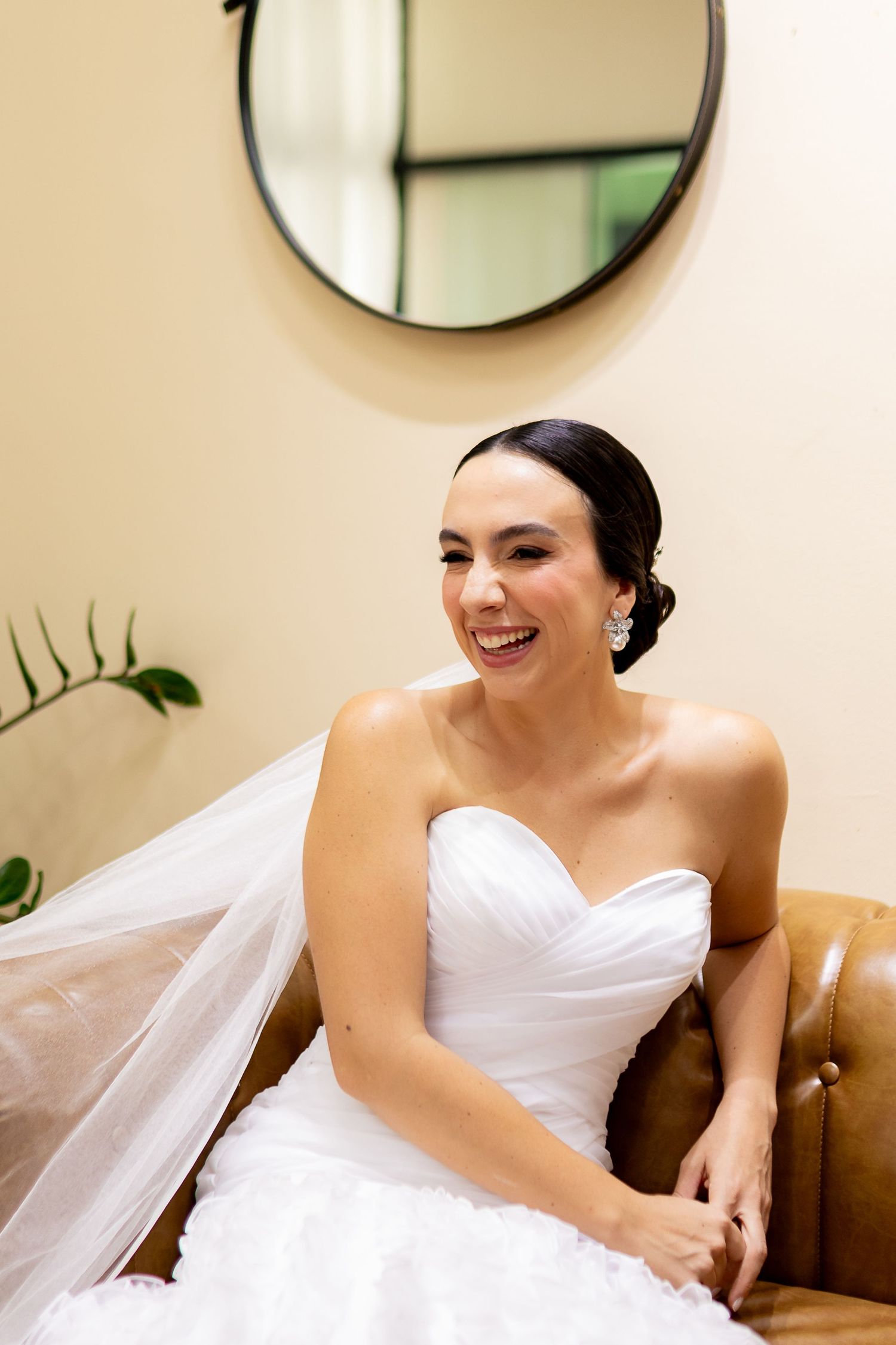 Smiling bride in white strapless gown with veil seated on leather sofa beneath round mirror.