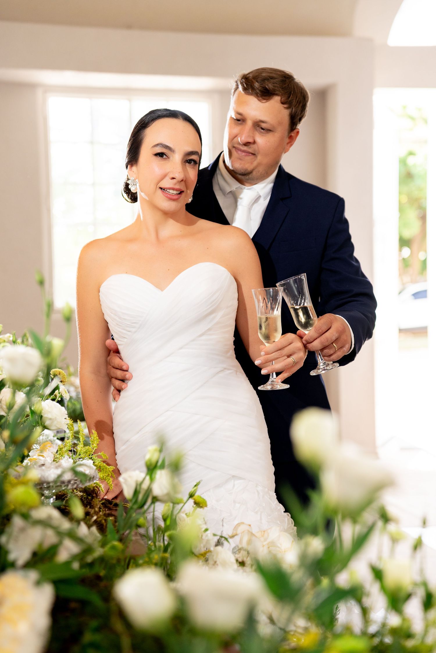Bride in white strapless gown and groom in navy suit holding champagne glasses among white floral arrangements.