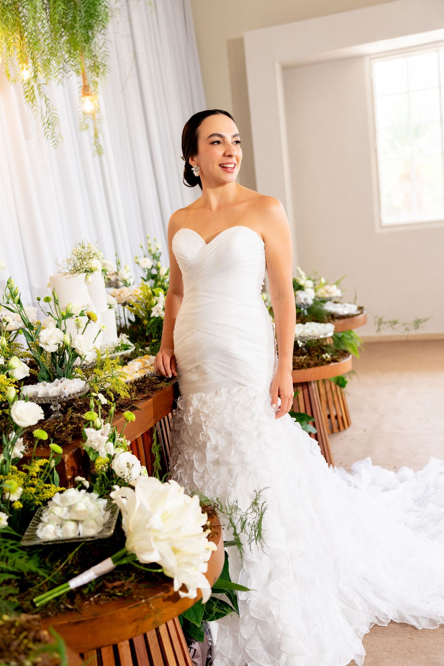 Smiling bride in a strapless mermaid wedding gown poses among lush white floral arrangements in an elegant indoor setting.