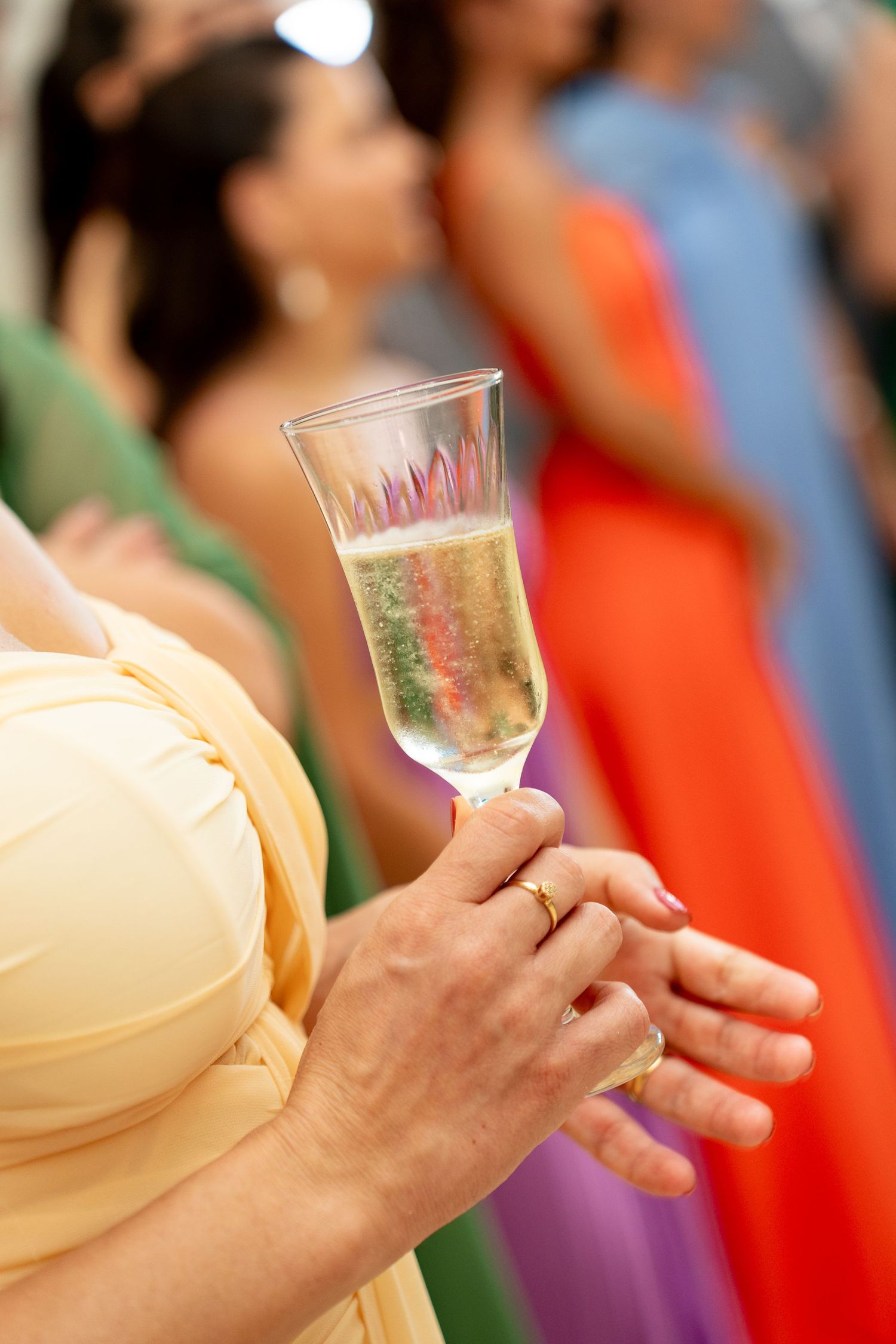 Hands holding a champagne flute at a colorful wedding reception with bridesmaids in vibrant dresses.