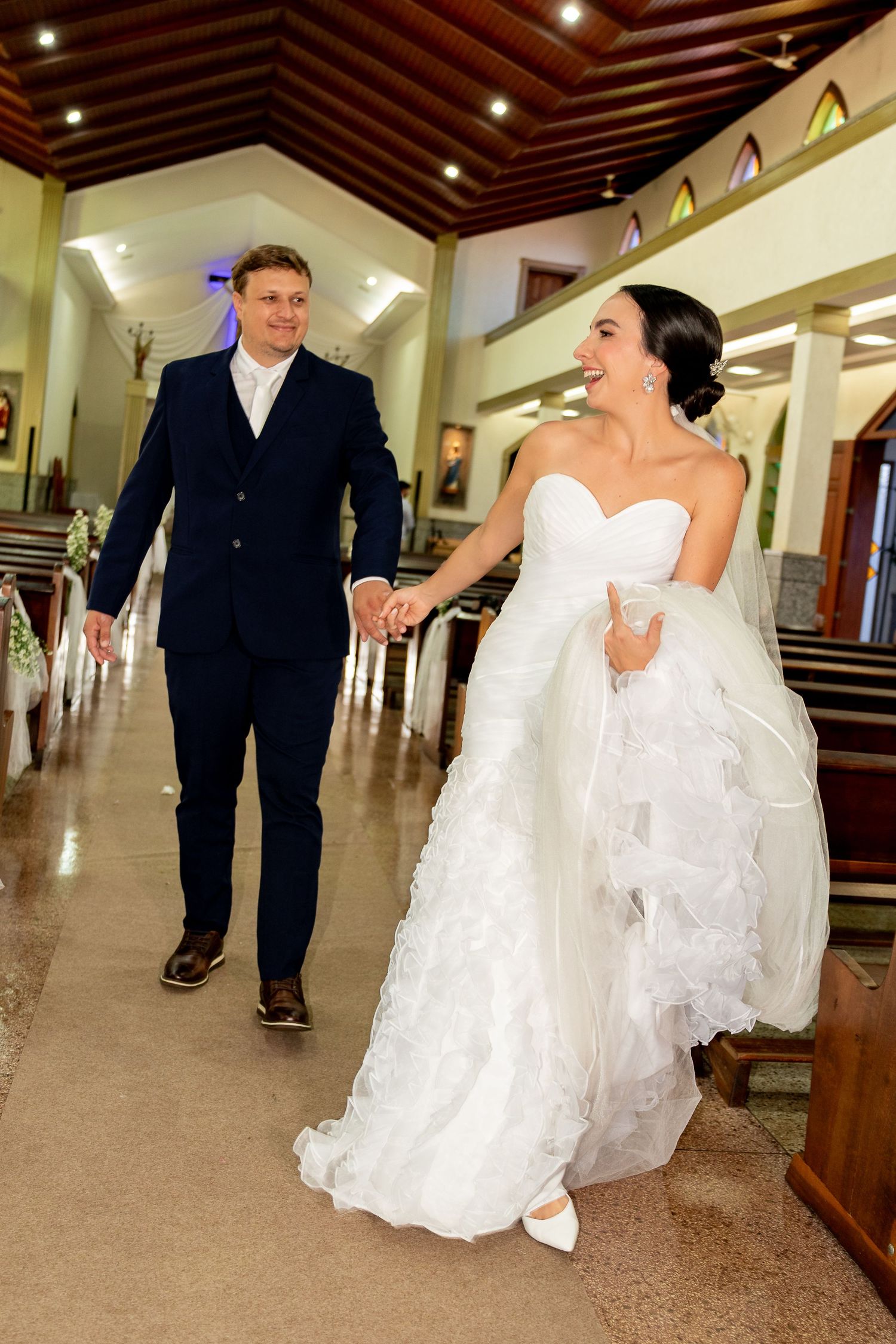 Bride and groom walk hand in hand down church aisle, she in white gown, he in navy suit, smiling joyfully.