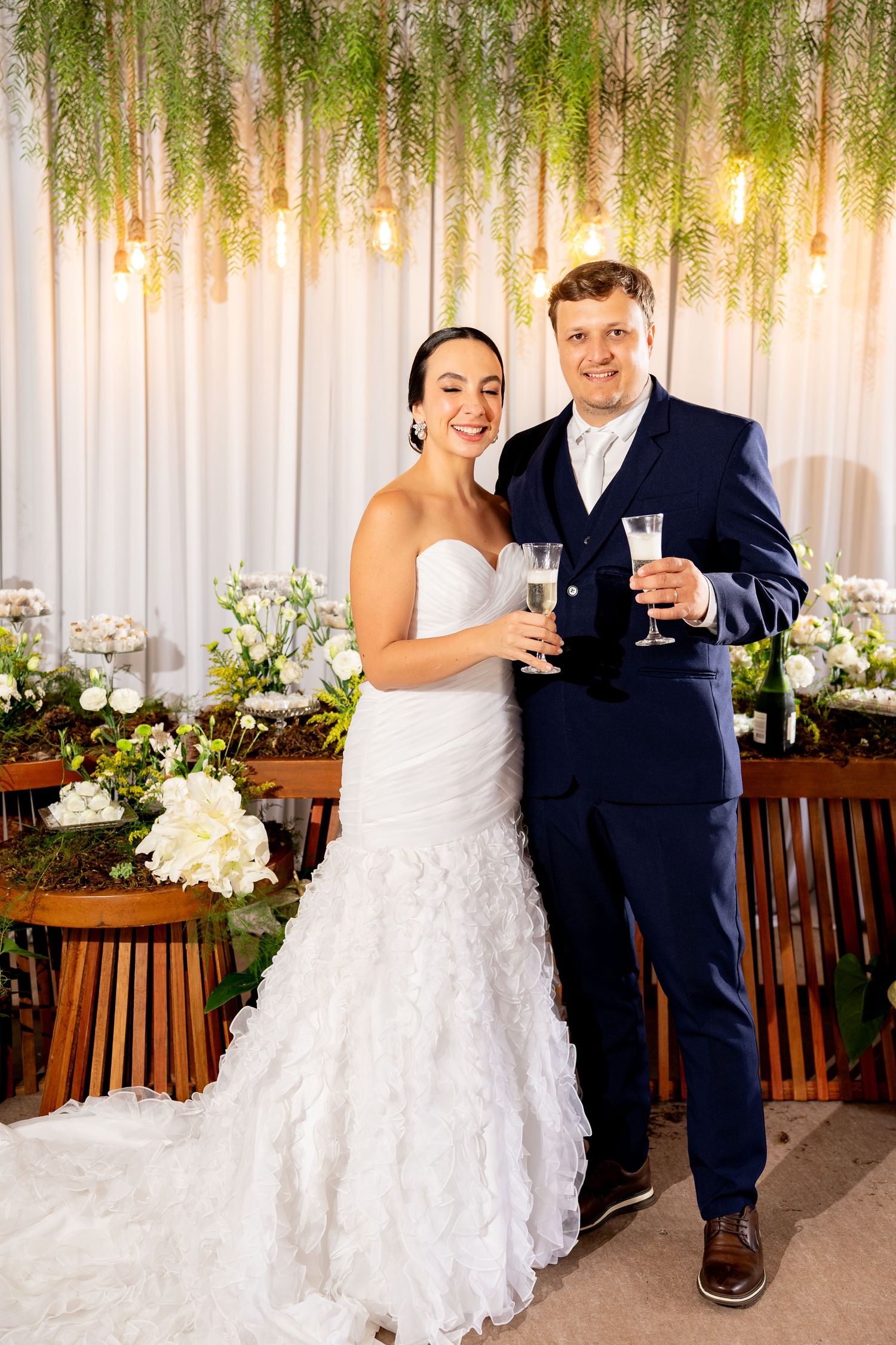 Bride in white ruffled gown and groom in navy suit toast with champagne at elegant floral wedding reception.