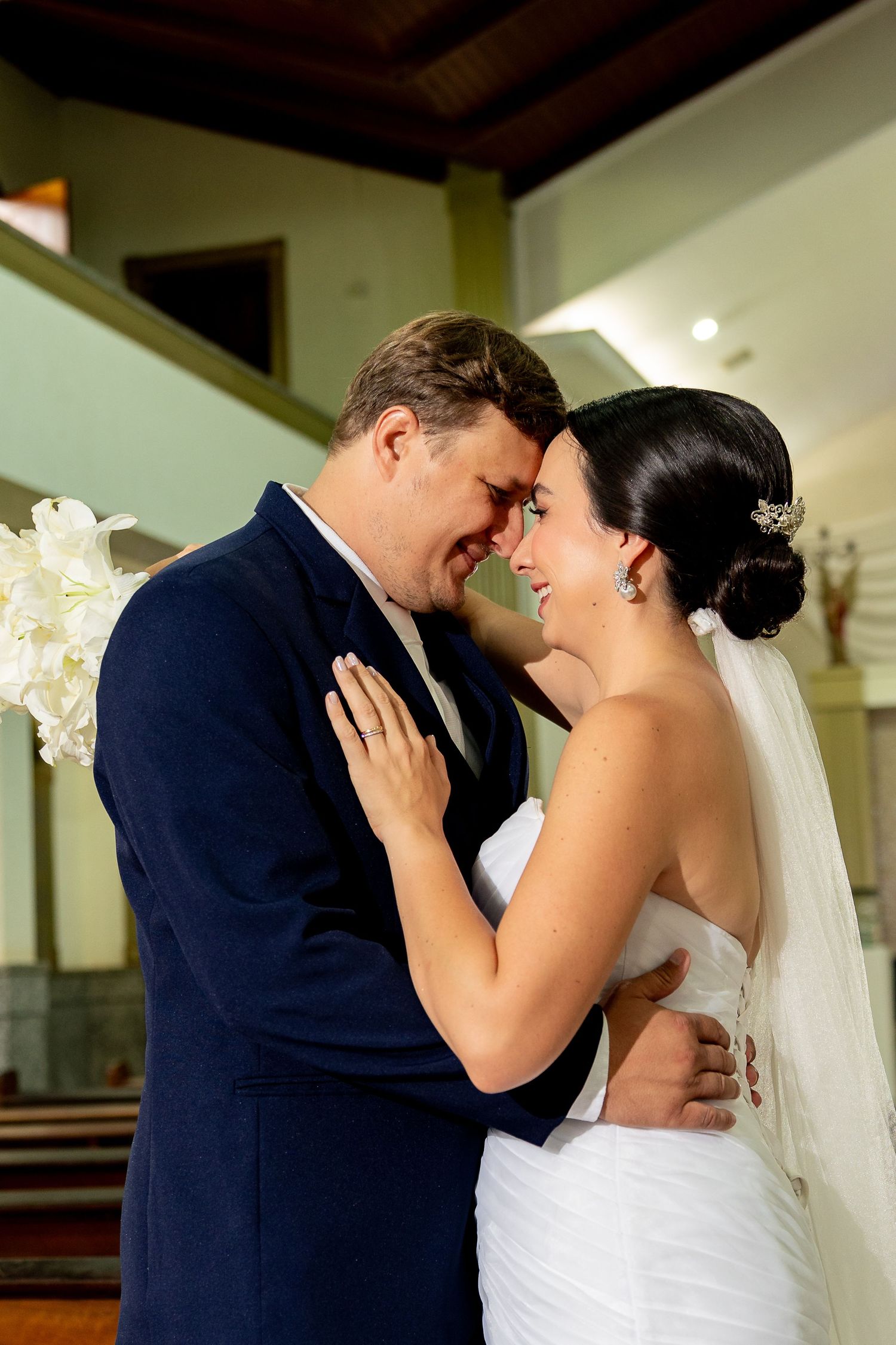 A bride and groom share an intimate moment, touching foreheads and smiling in a church on their wedding day.