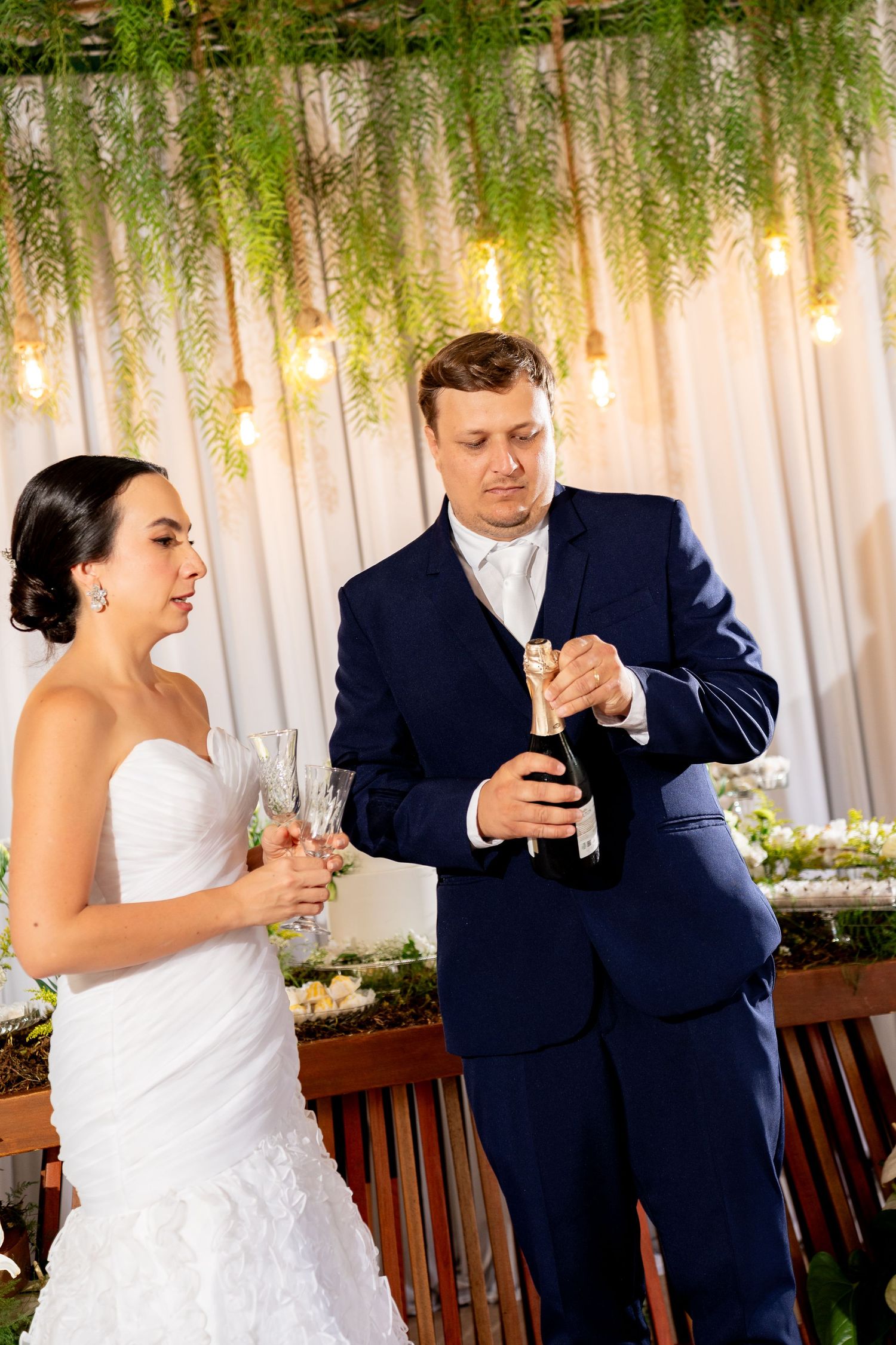 Bride in white gown holds champagne flutes as groom in navy suit opens a champagne bottle at wedding reception.