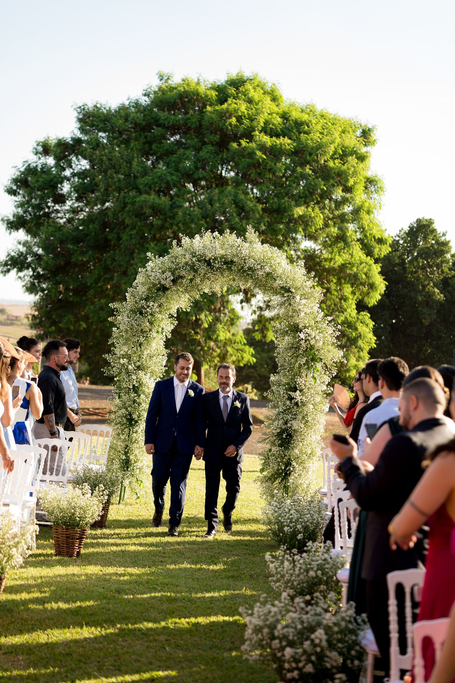 Two grooms walk down the aisle at an outdoor wedding ceremony beneath a floral arch surrounded by guests.
