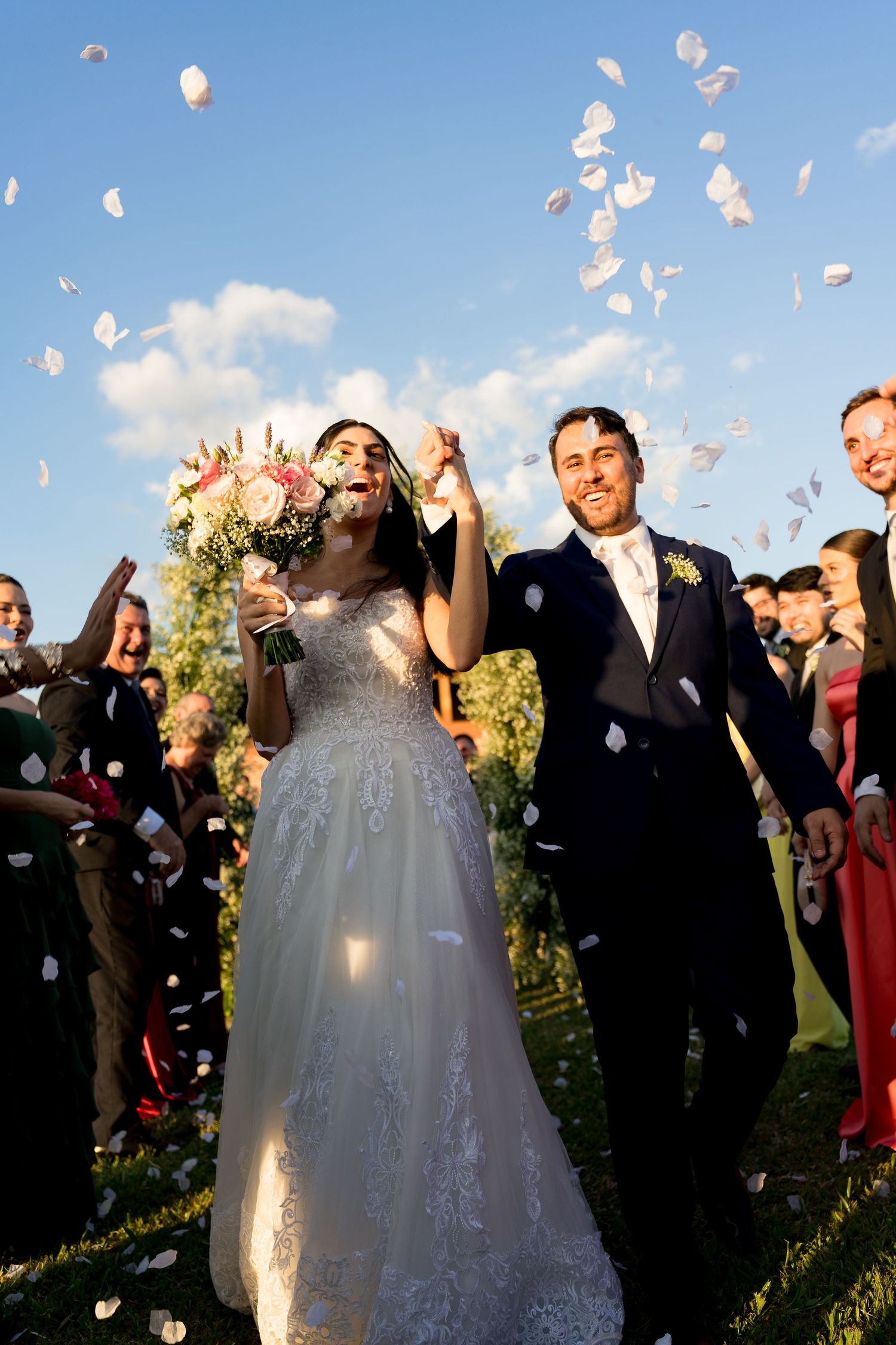 Joyful newlyweds walk through a shower of white petals surrounded by cheering guests under a blue sky.