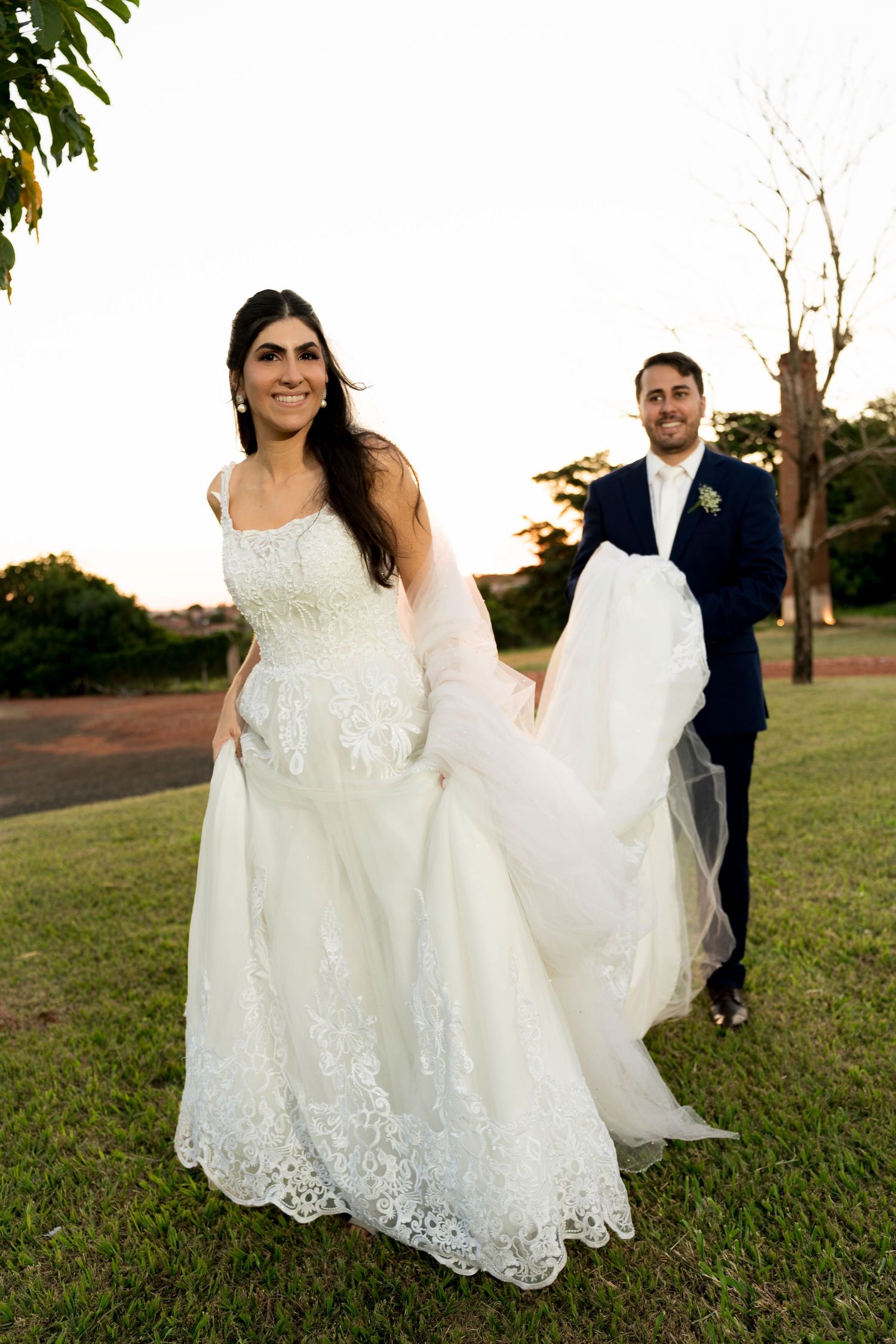 Bride in white lace gown walks outdoors as groom in navy suit carries her train at golden hour.