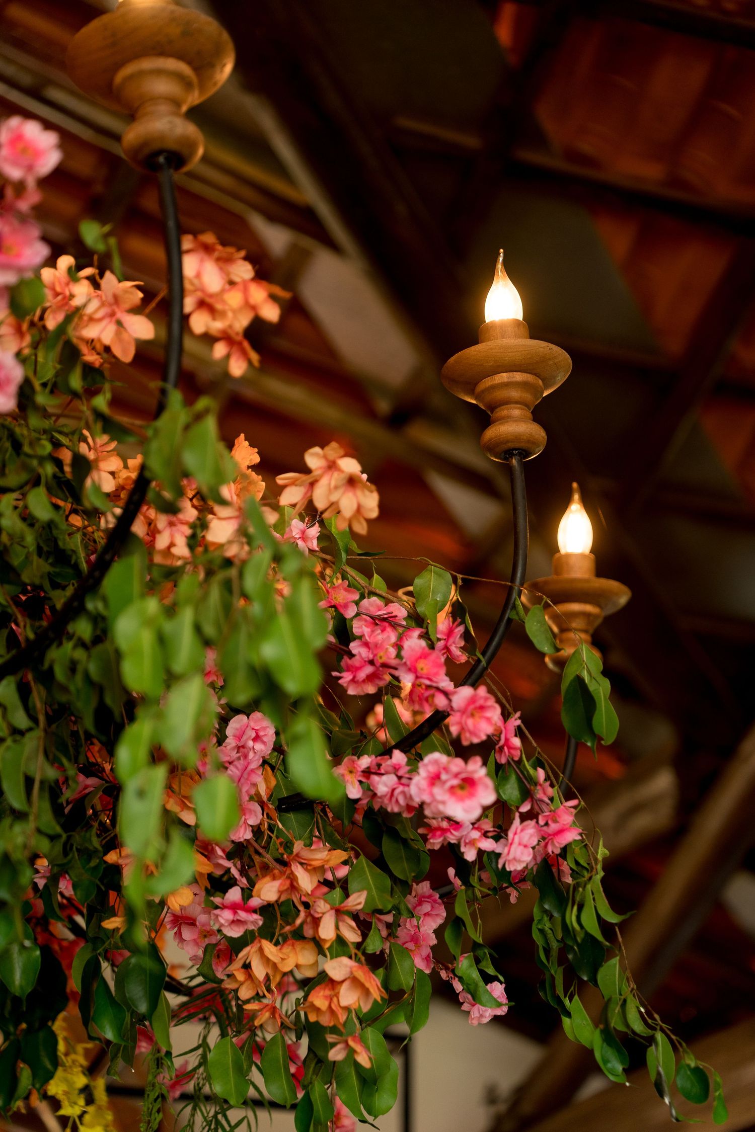 Wooden chandelier with lit candle bulbs adorned with cascading pink and green flowers hanging from a rustic ceiling.