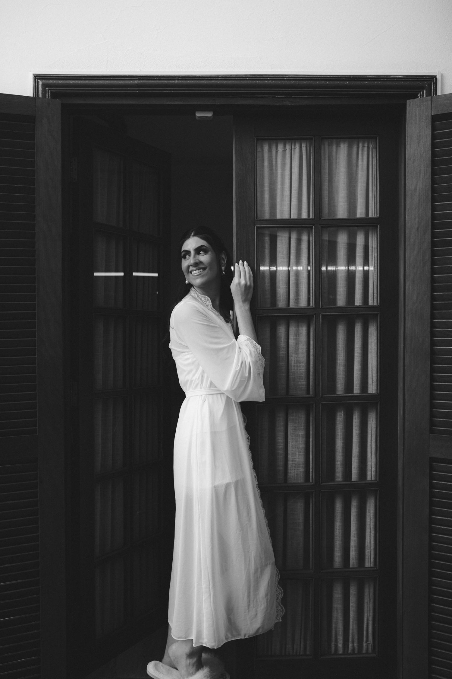 Black and white photo of a woman in a white dress standing in an open doorway, smiling and looking up.
