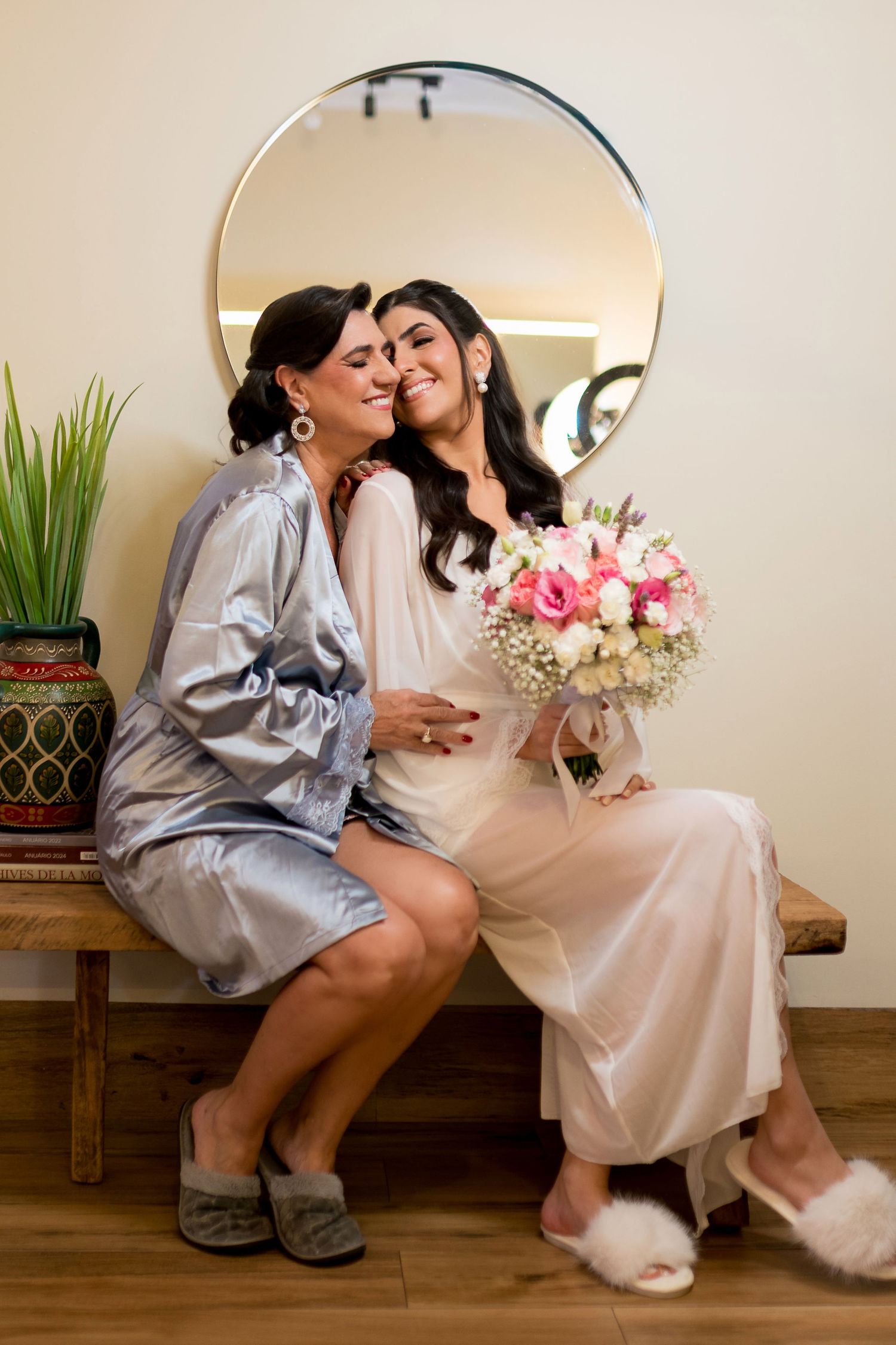 Bride in white robe holding pink bouquet sits with woman in silver satin robe, sharing a tender moment before a round mirror.