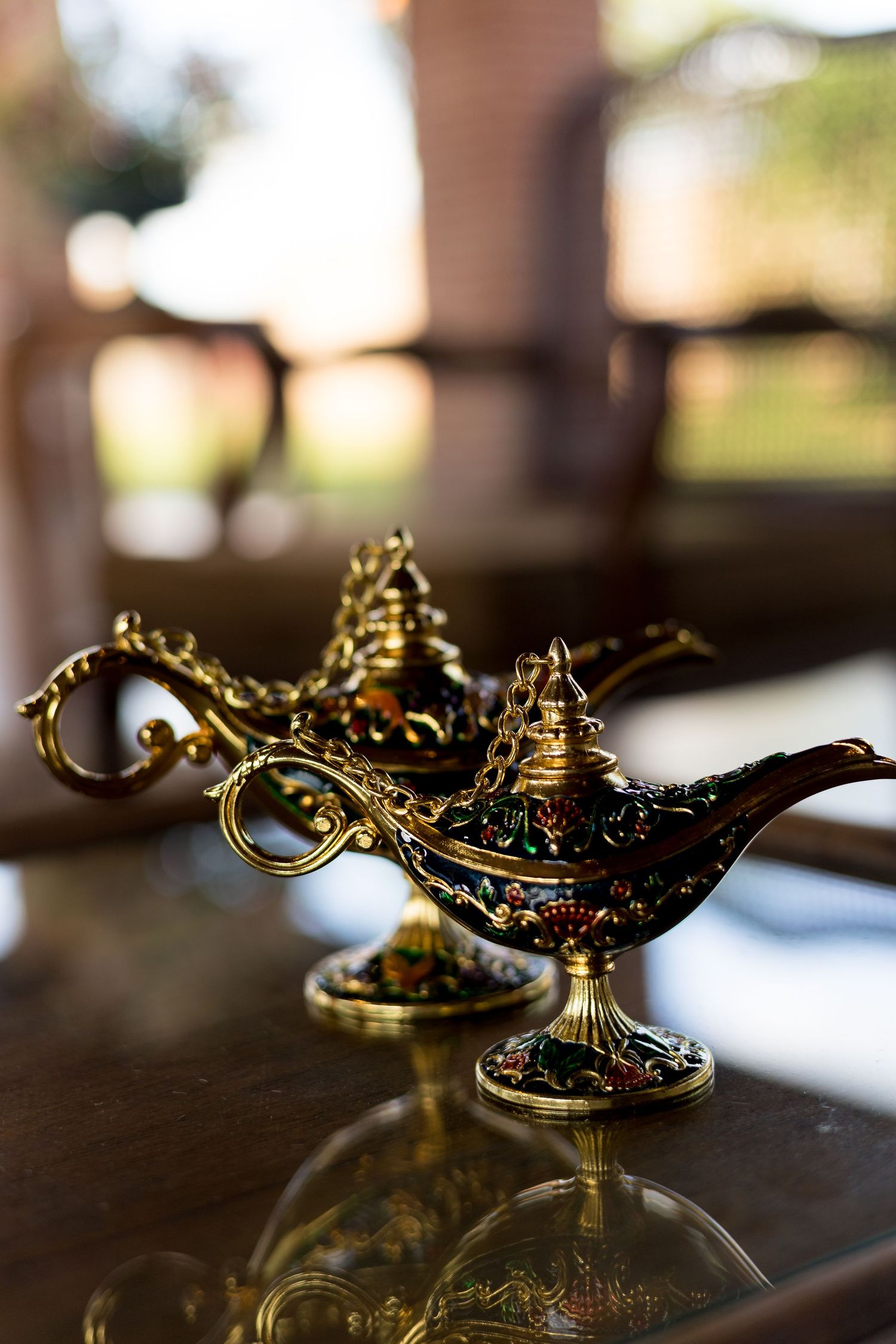 Two ornate golden Aladdin genie lamps with intricate detailing sit on a reflective glass surface.