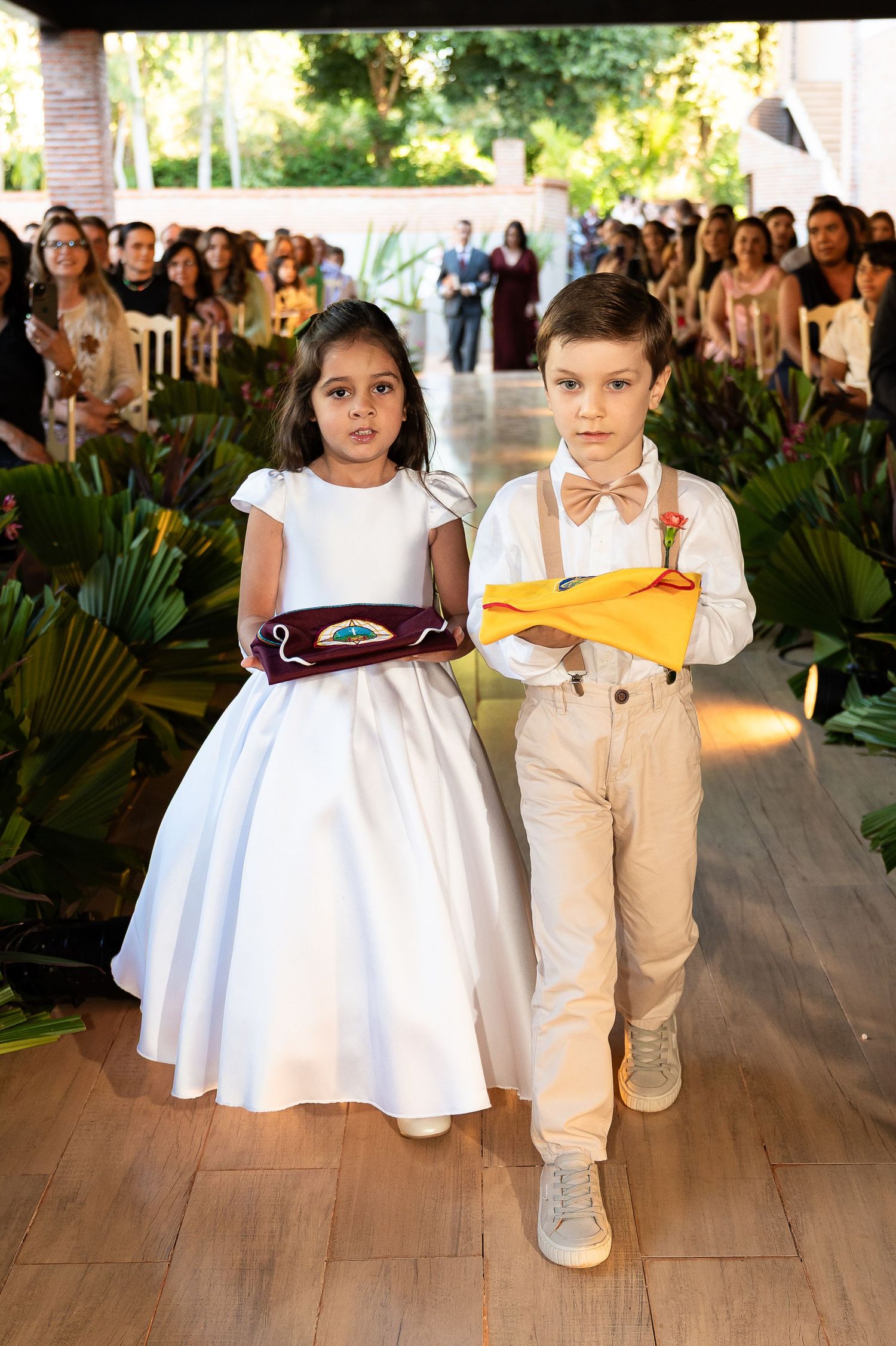 Two children in formal attire walk down a wedding aisle carrying ring pillows at an outdoor ceremony.