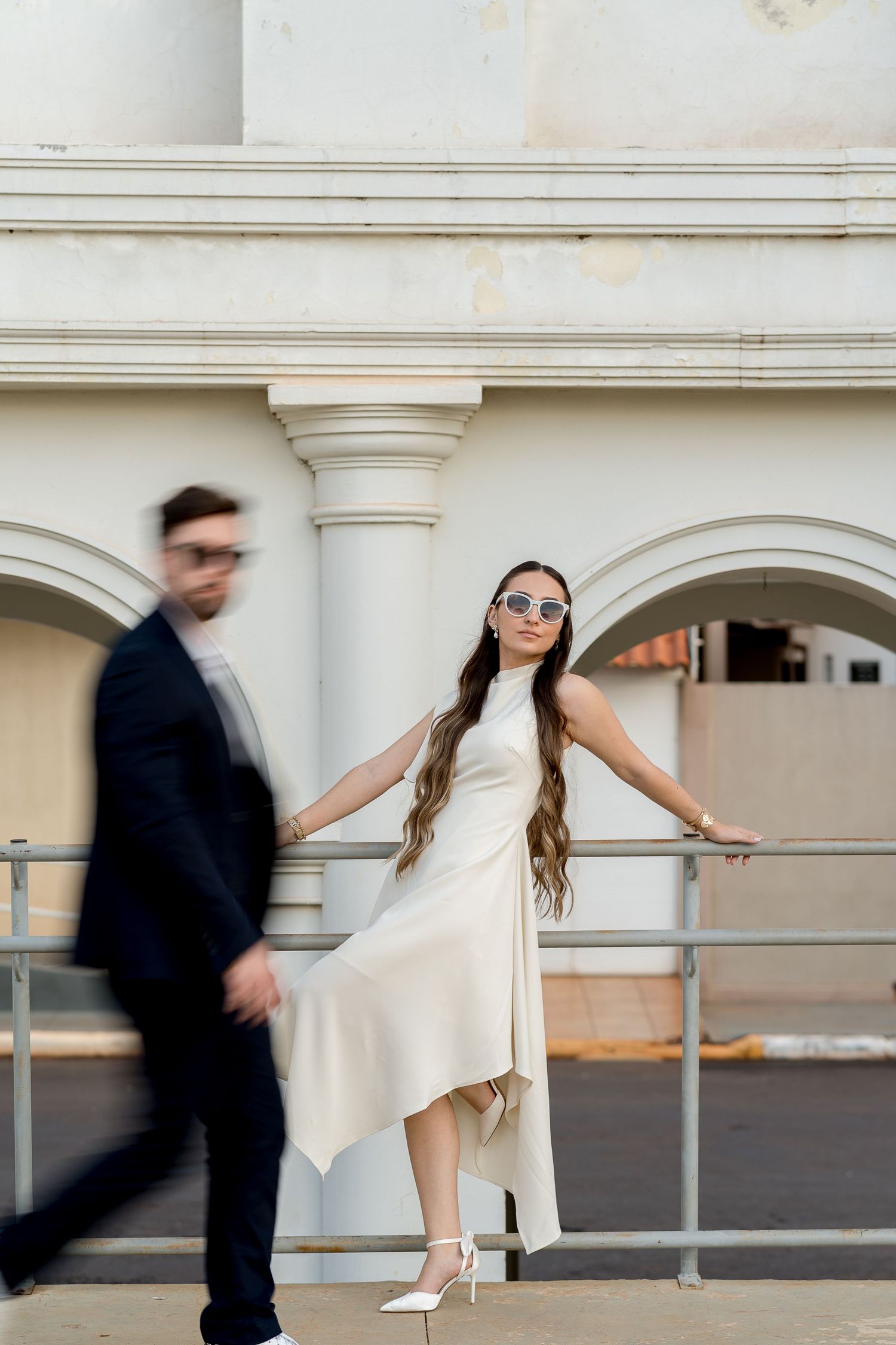 Elegant woman in white dress and sunglasses poses by white columns while man in suit walks past.