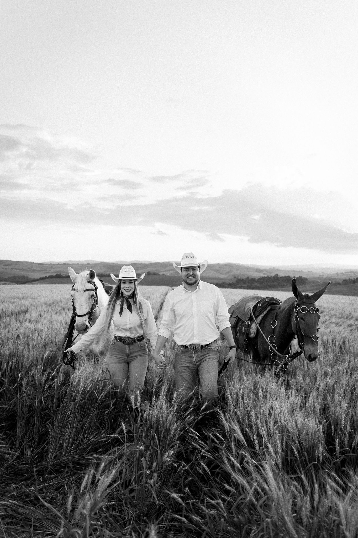 Black and white photo of a couple in cowboy hats walking with two horses through a wheat field.
