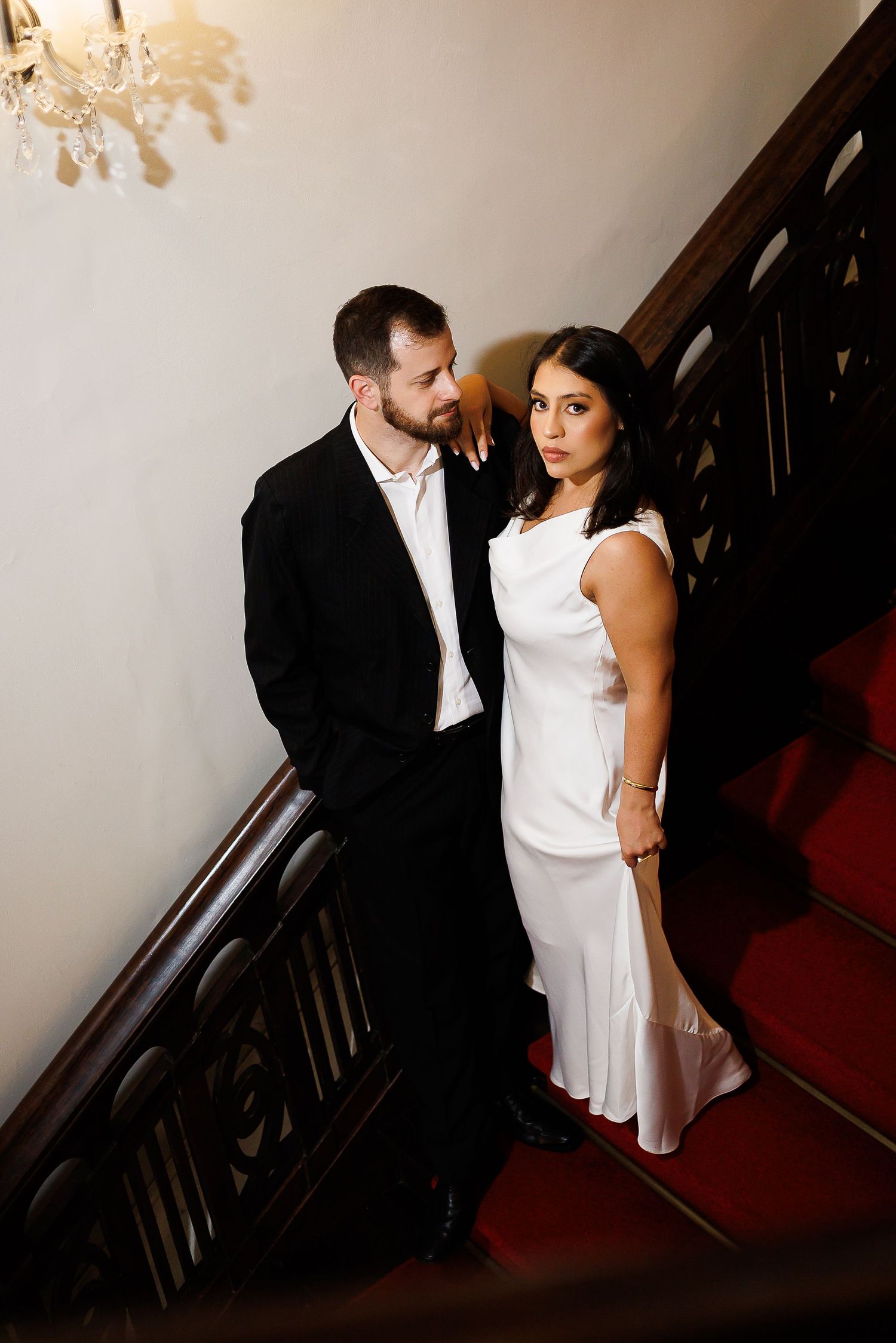 Elegant couple in formal attire posing on a grand staircase with red carpet and dark wood railing.