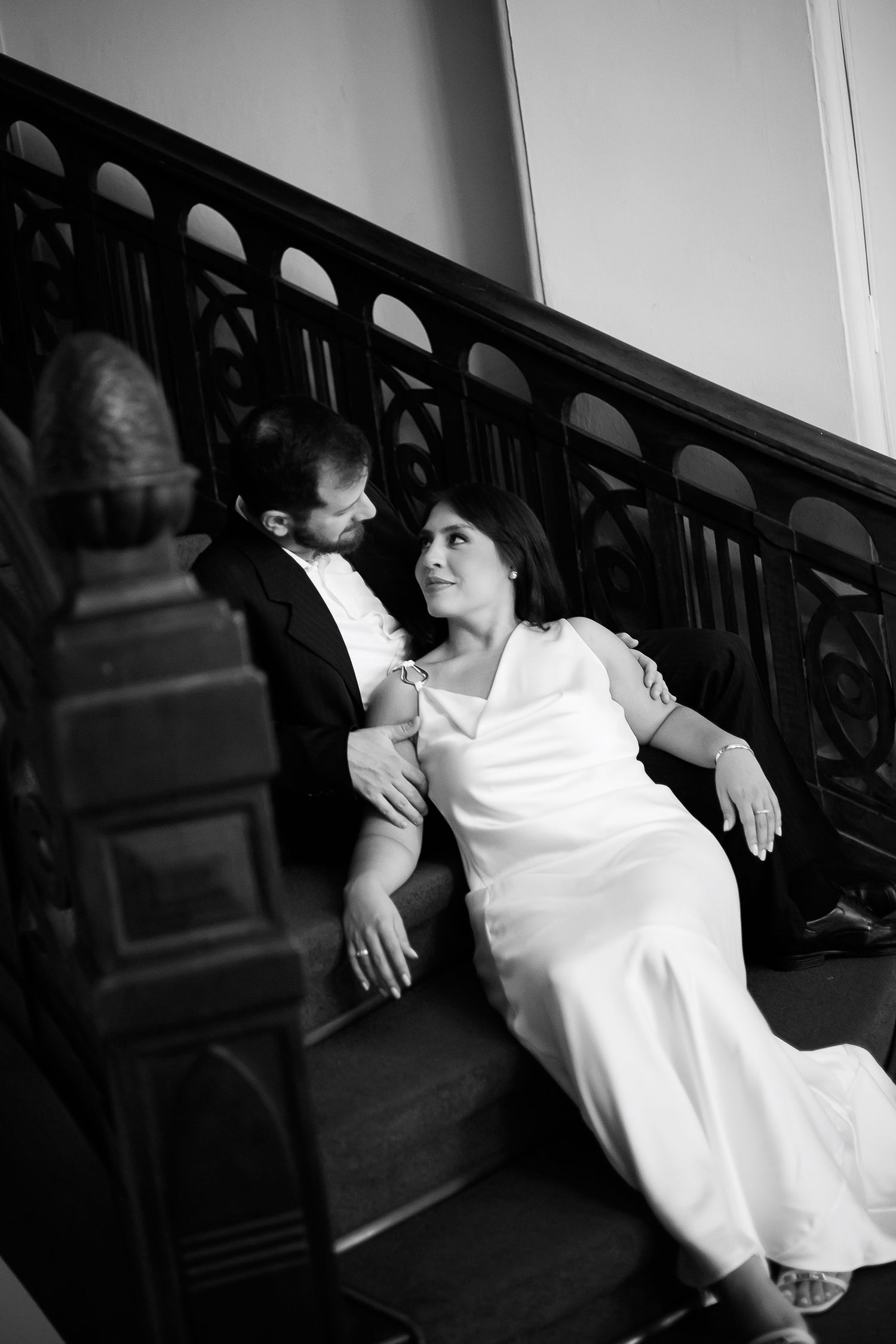 Black and white photo of a couple in wedding attire sitting on ornate staircase, gazing at each other romantically.