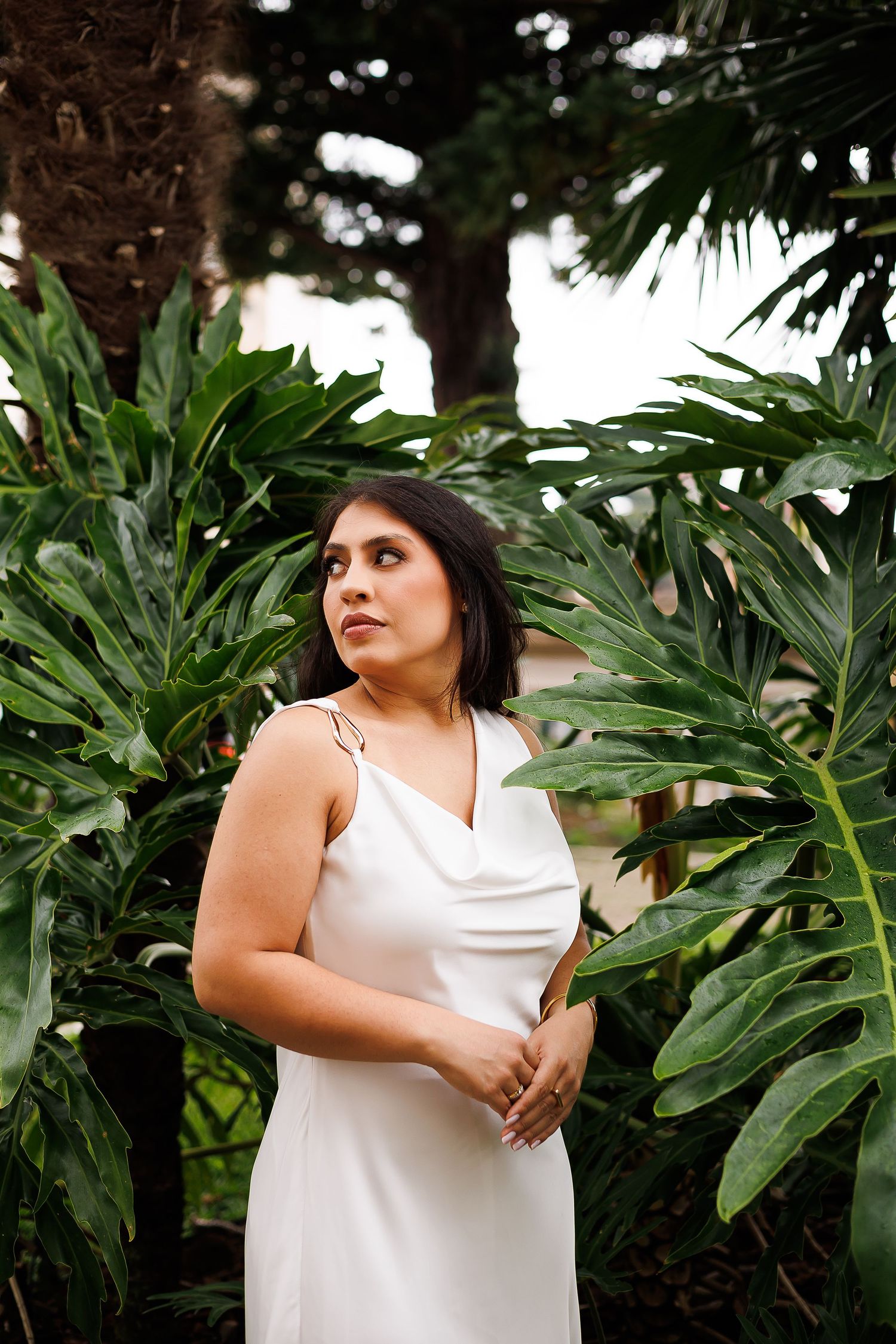 Woman in white satin slip dress posing among tropical monstera leaves and palm trees in a lush garden setting.