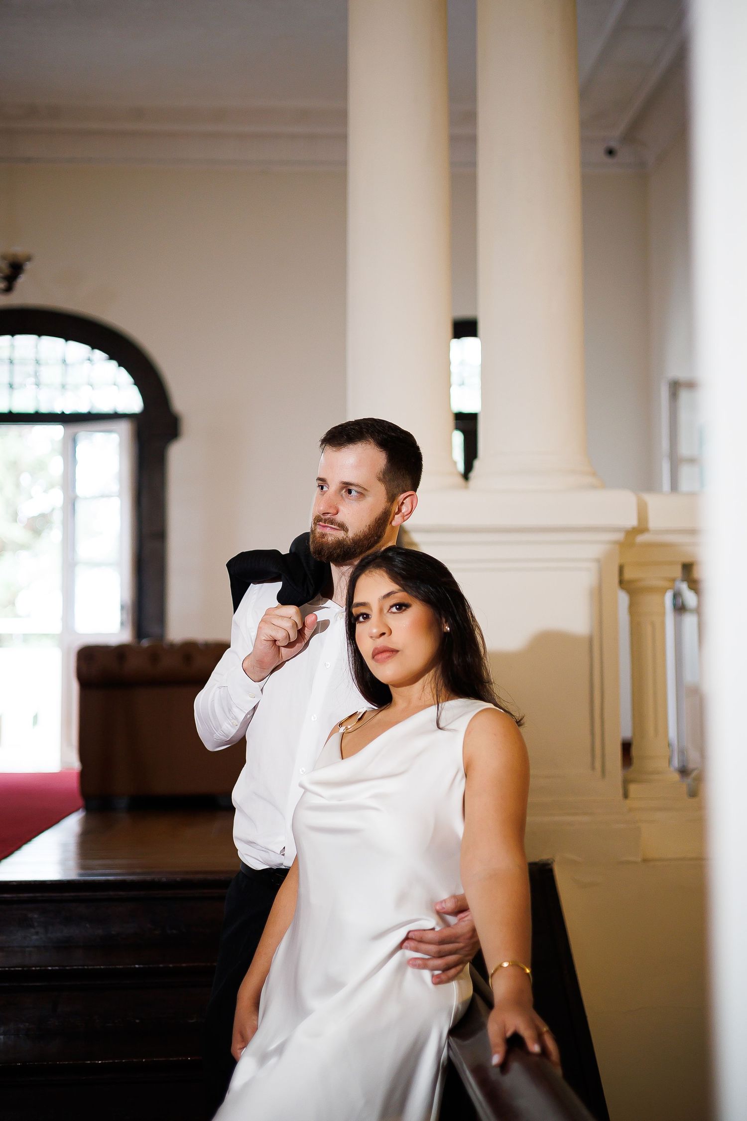 Elegant couple in wedding attire posing inside a grand venue with white columns, man in suit embracing woman in white gown.