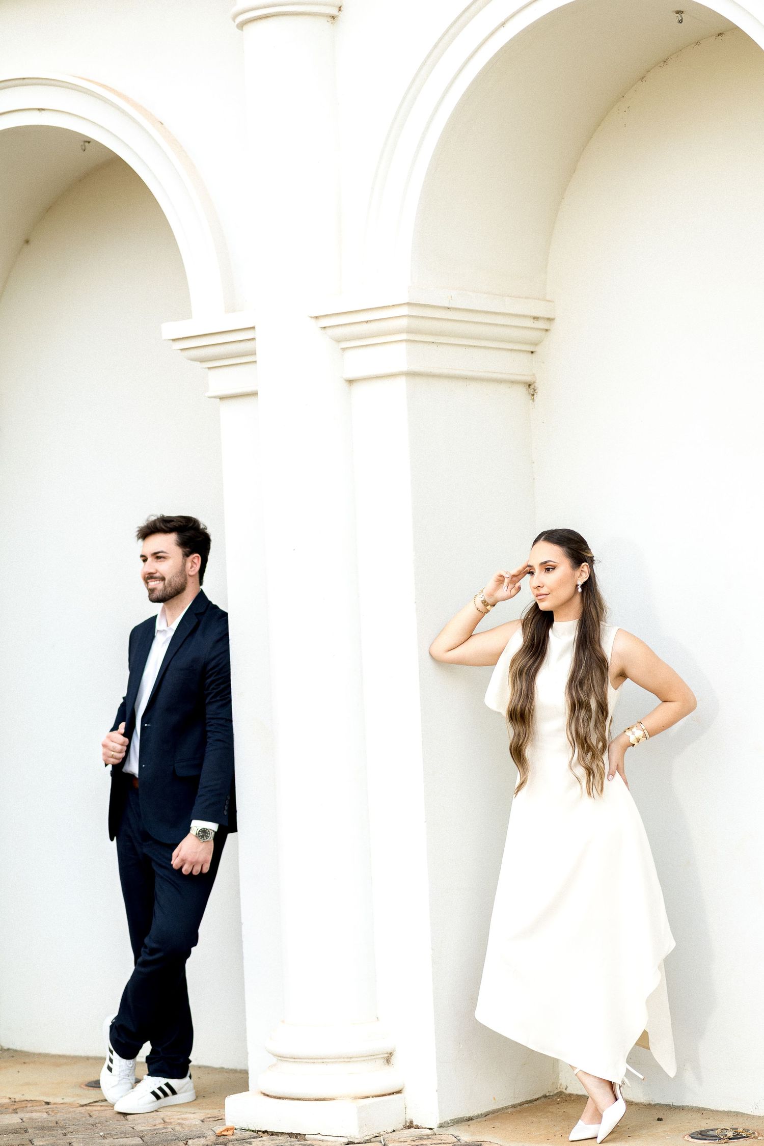 Elegant couple posing by white arched columns, man in black suit, woman in flowing white dress.