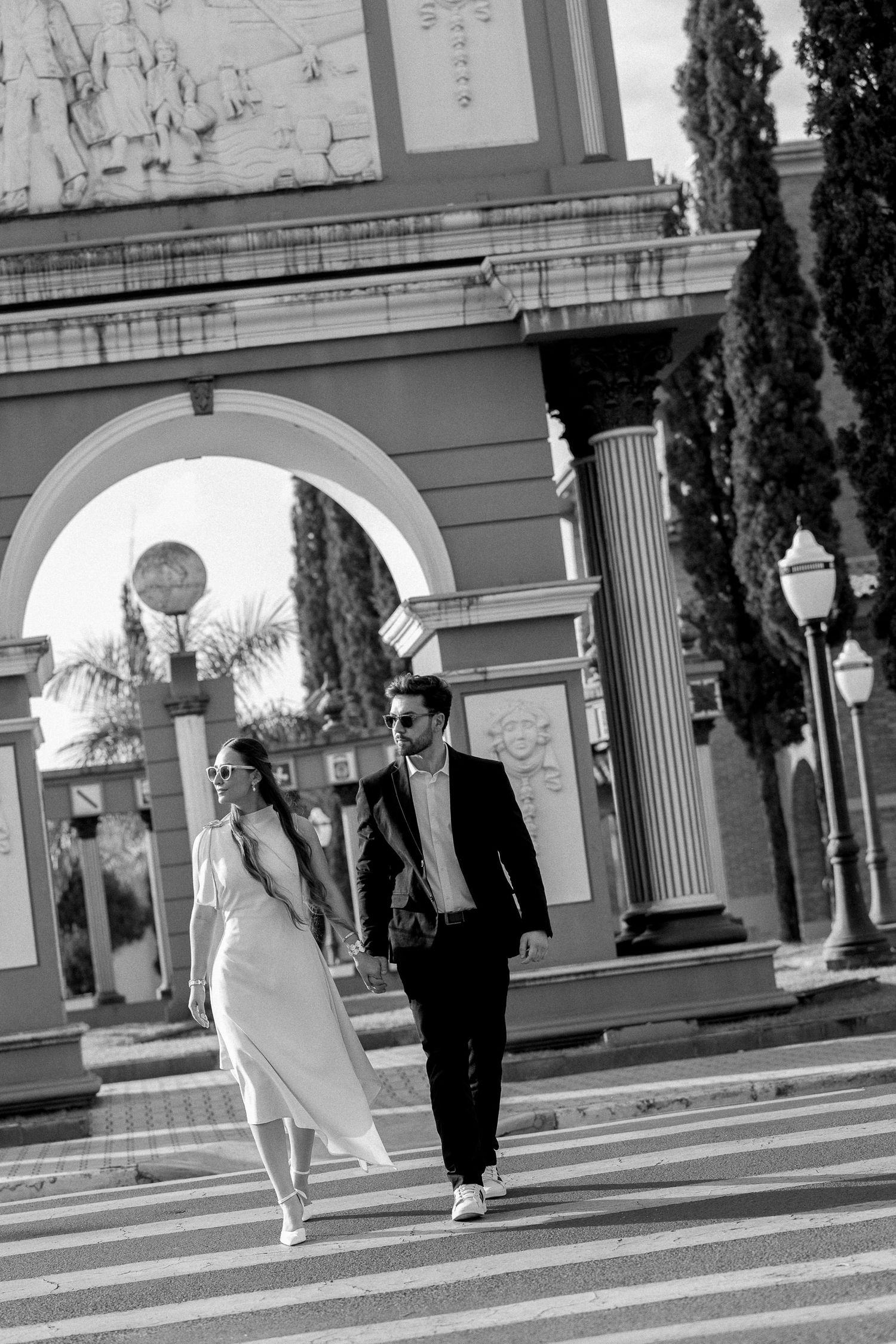 Black and white photo of a stylish couple holding hands while crossing a street near a grand archway with columns.