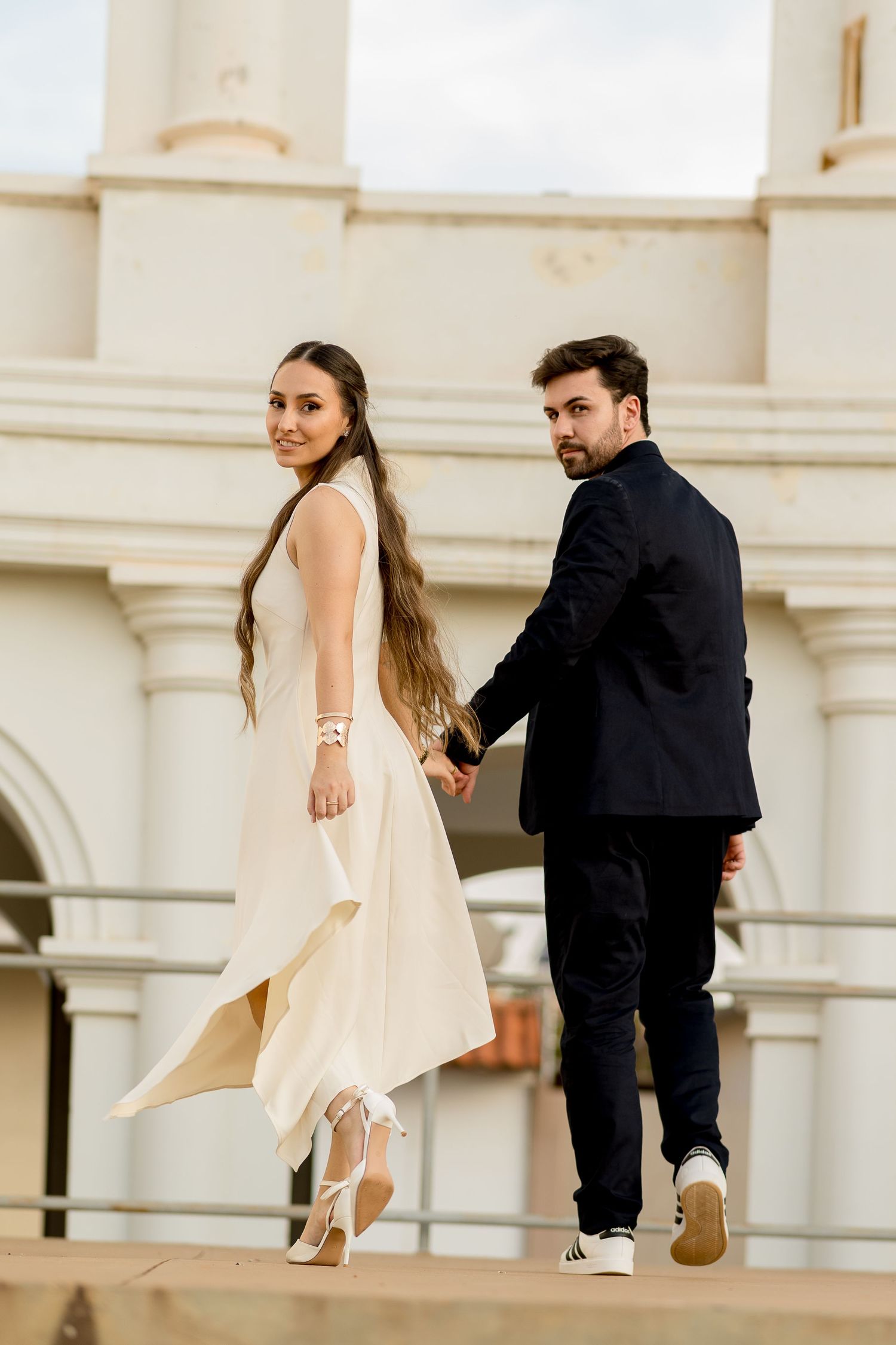 Elegant couple in formal wear holding hands outside a white classical building, bride in flowing white dress.