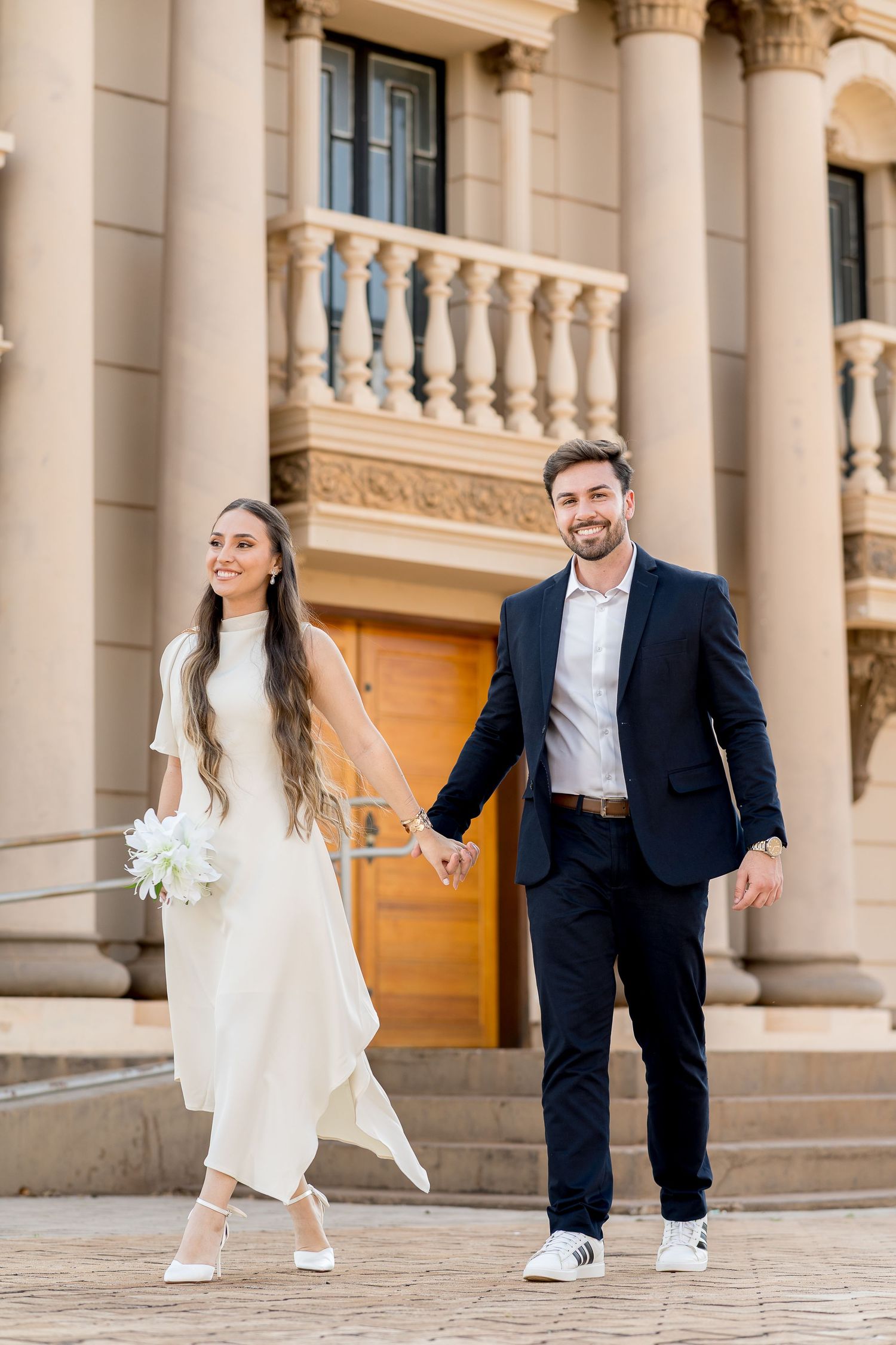 Smiling bride and groom holding hands walking outside elegant building, bride in white dress holding bouquet, groom in navy suit.