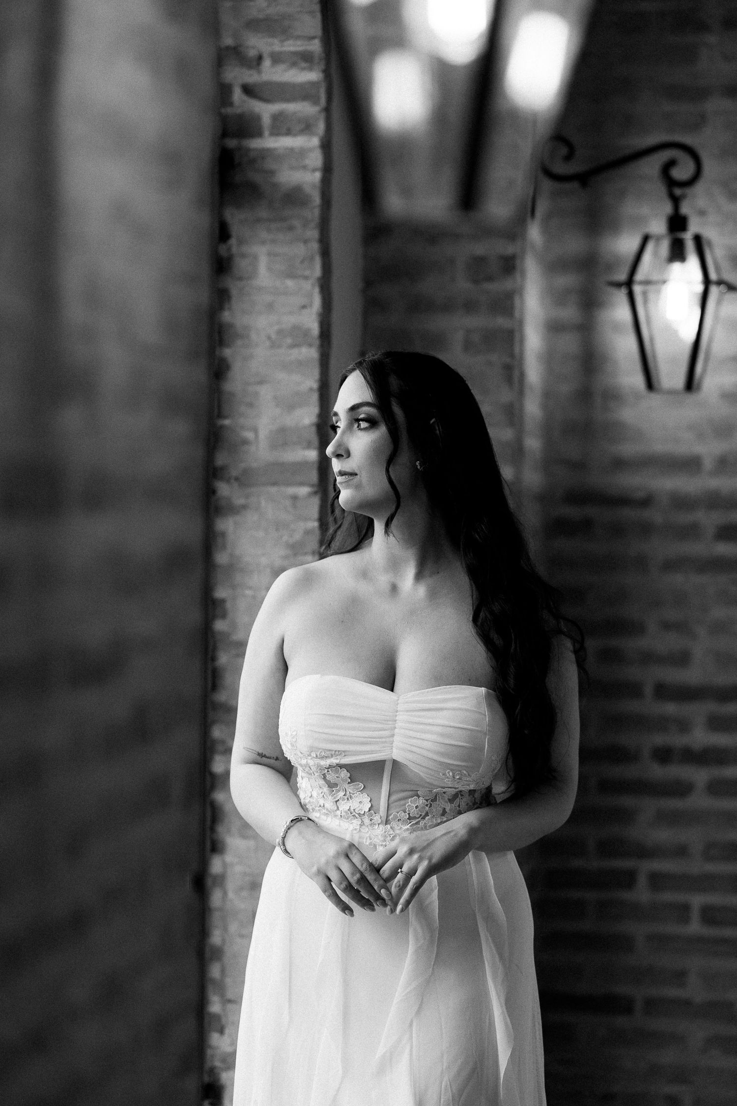 Black and white photo of a bride in a strapless wedding gown standing by a brick wall with a vintage lantern.