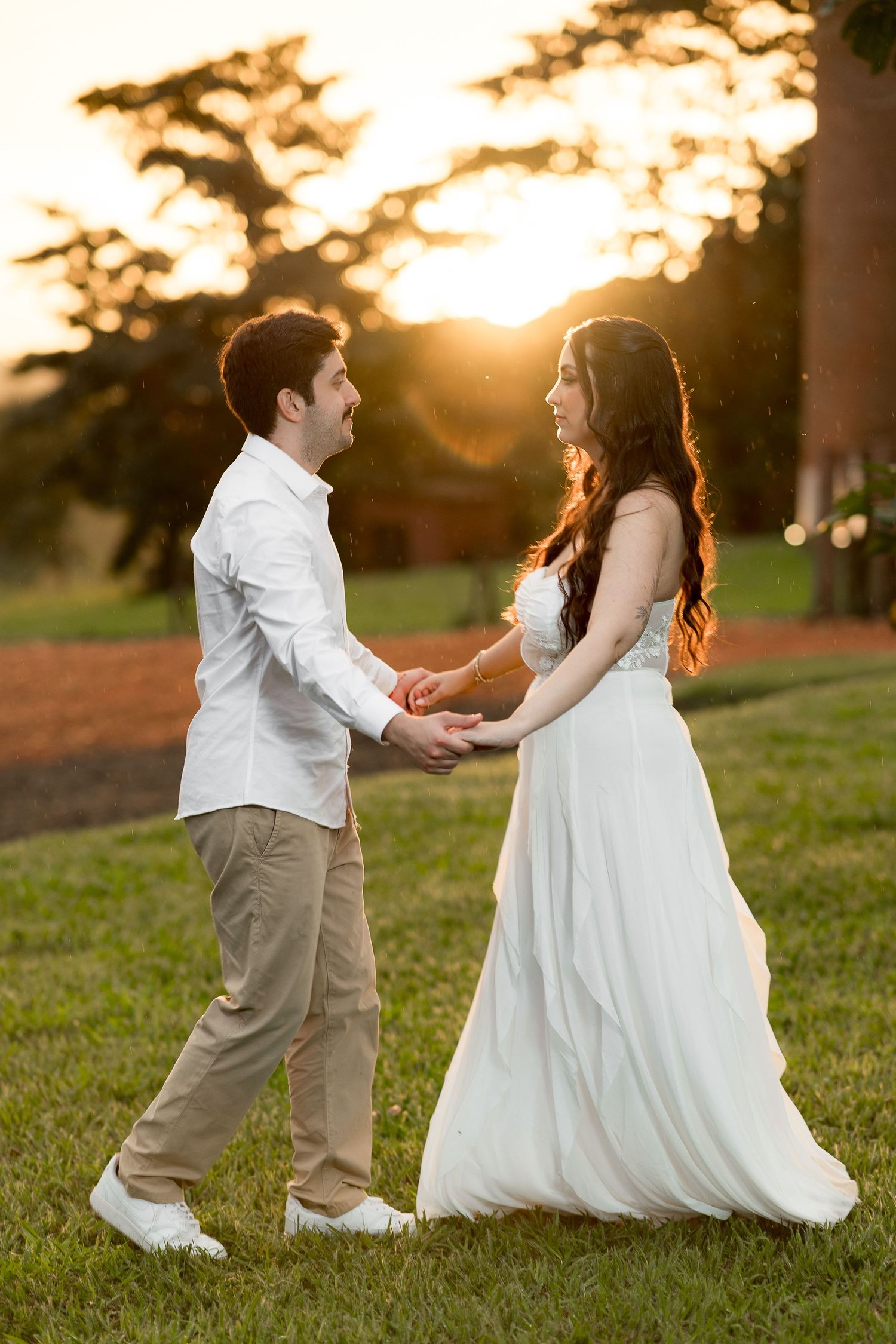 Couple holding hands in white wedding attire at golden hour sunset in a lush green park.