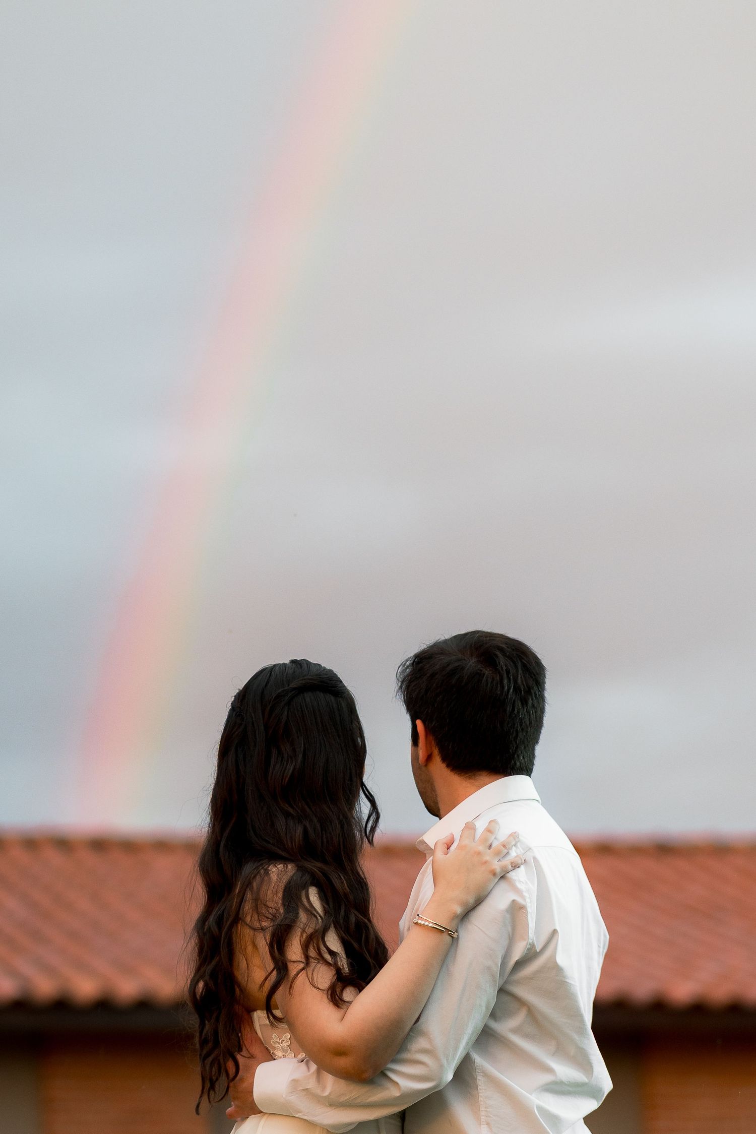 Couple embracing while gazing at a vibrant rainbow in a cloudy sky, viewed from behind, romantic outdoor moment.