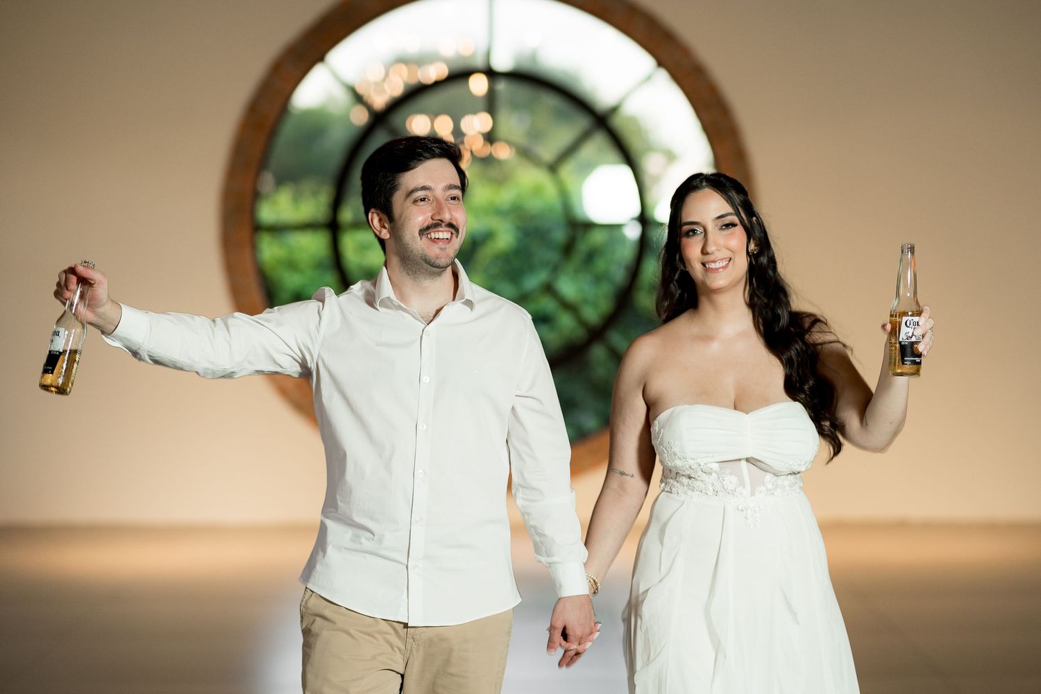 A smiling couple in white attire toasting with beer bottles in an elegant venue with a round window.