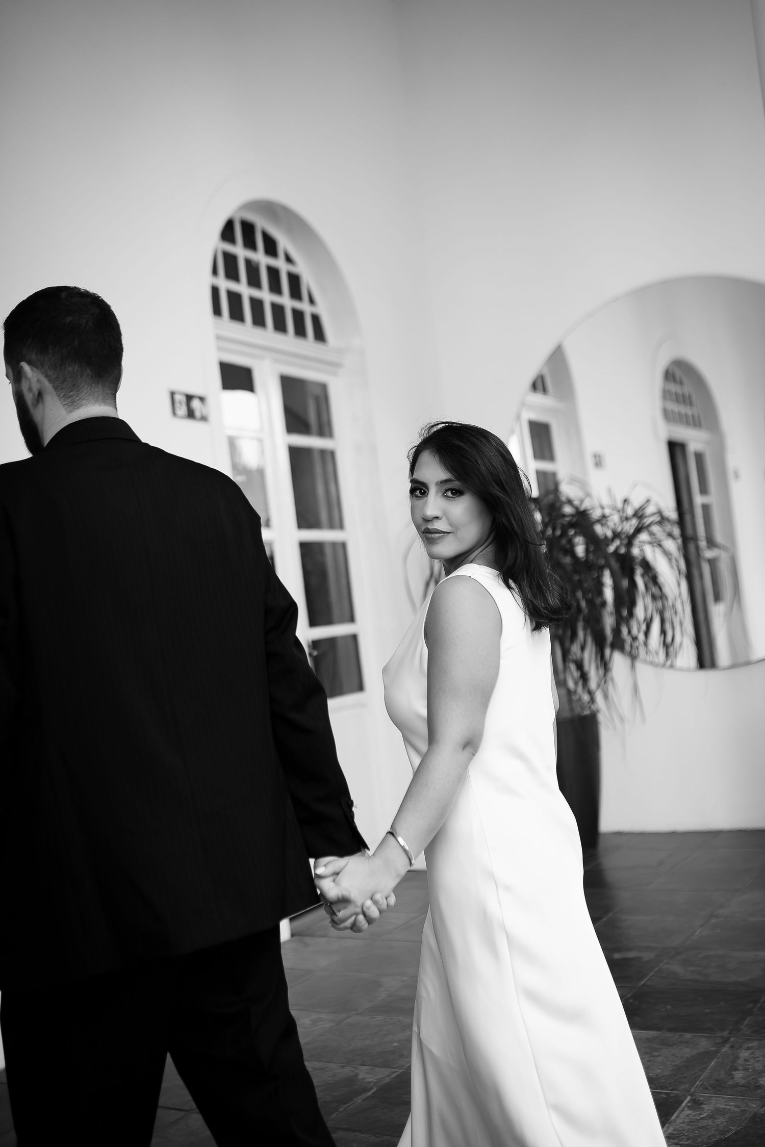 Black and white photo of a bride in white dress holding hands with groom in black suit, looking back at camera.