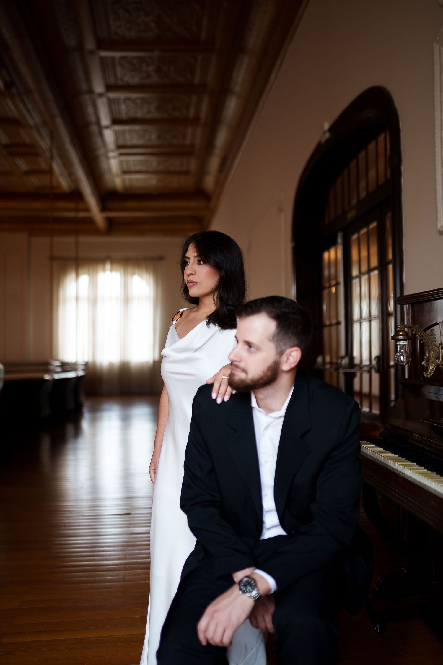 Elegant couple in formal wear posing near a vintage piano in a grand historic hall with ornate ceiling.
