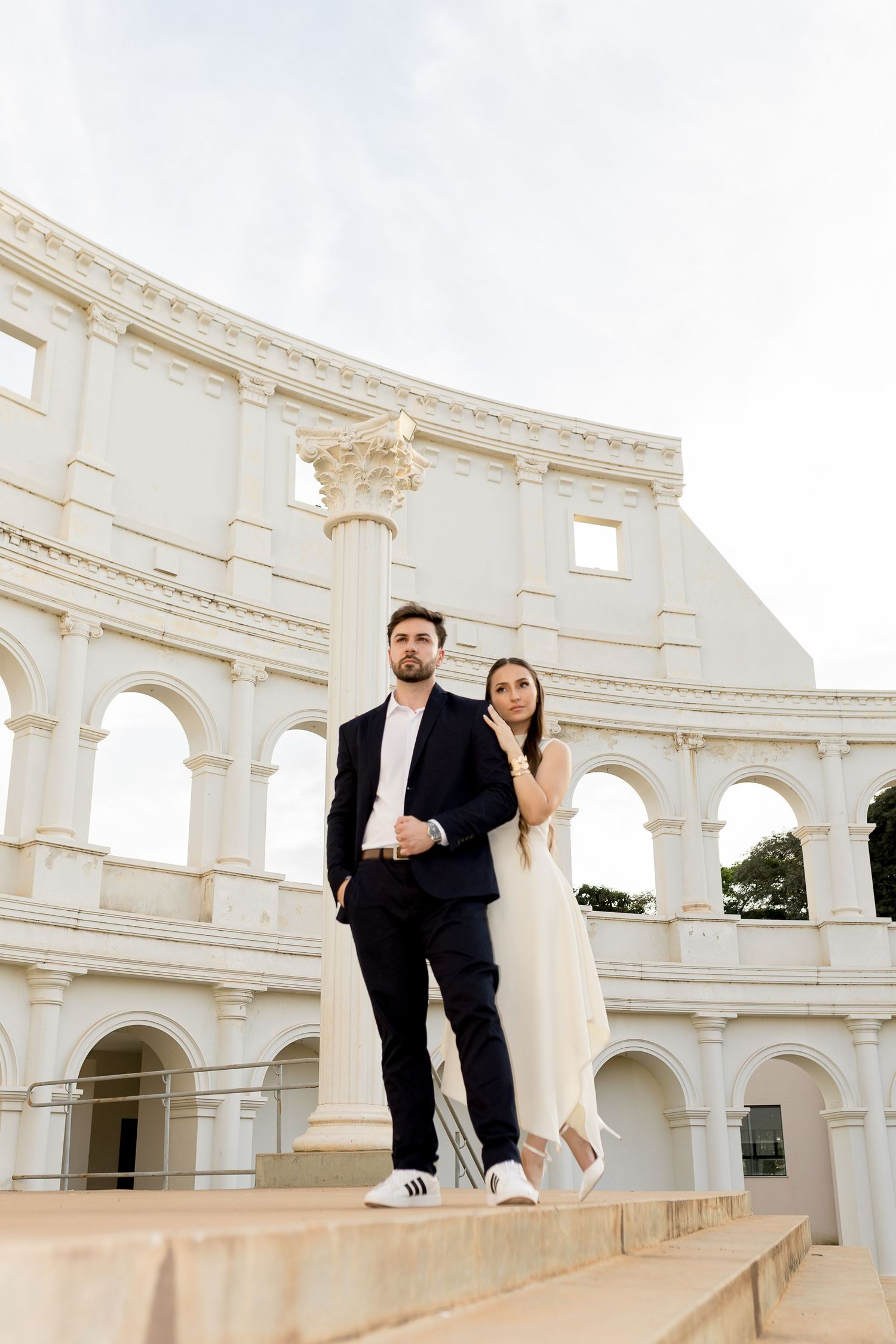 Elegant couple posing on steps before a white Roman Colosseum-inspired architecture, man in suit, woman in white dress.