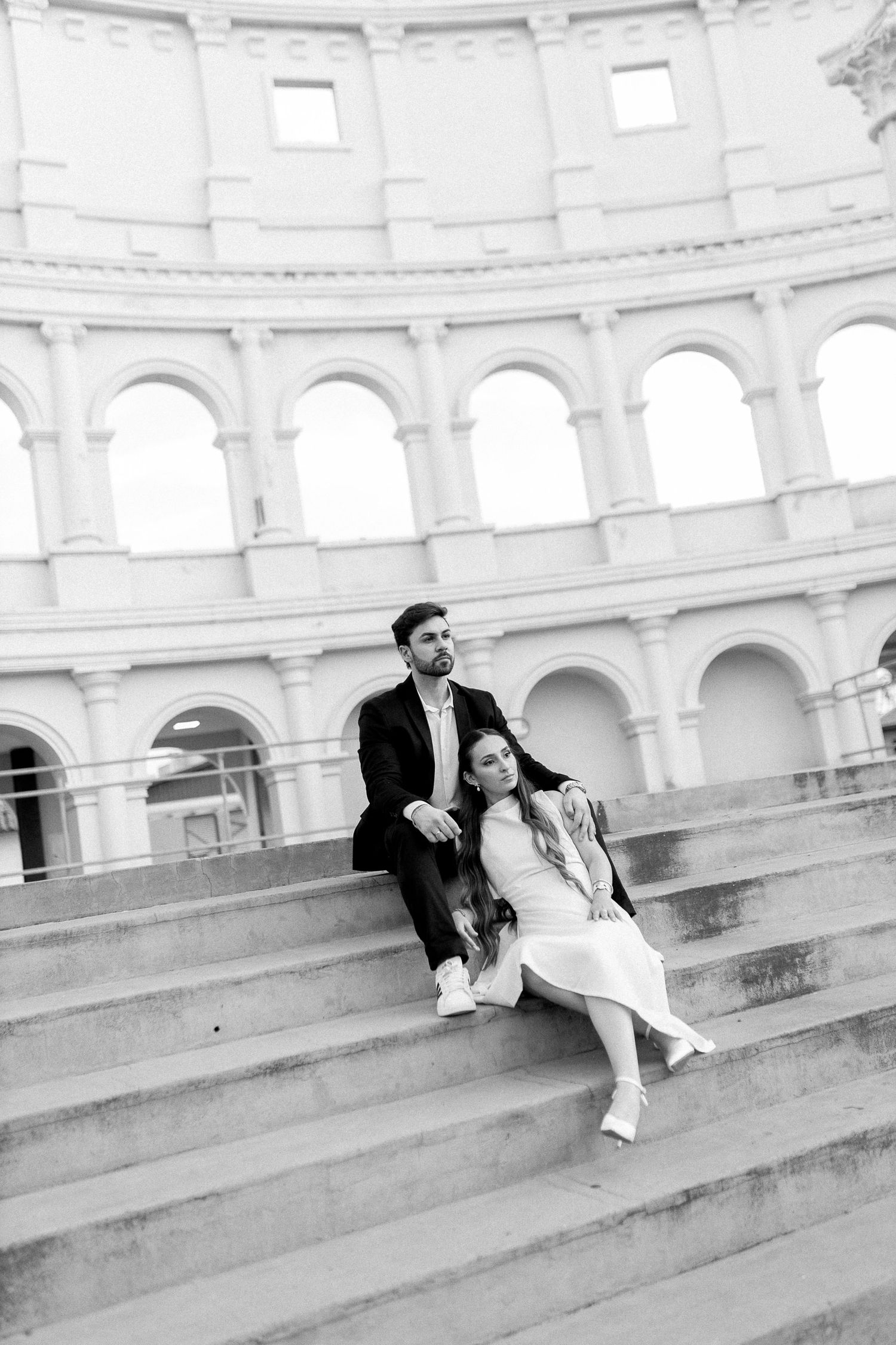 Black and white photo of a couple sitting on steps in front of a grand arched building, sharing a romantic moment.