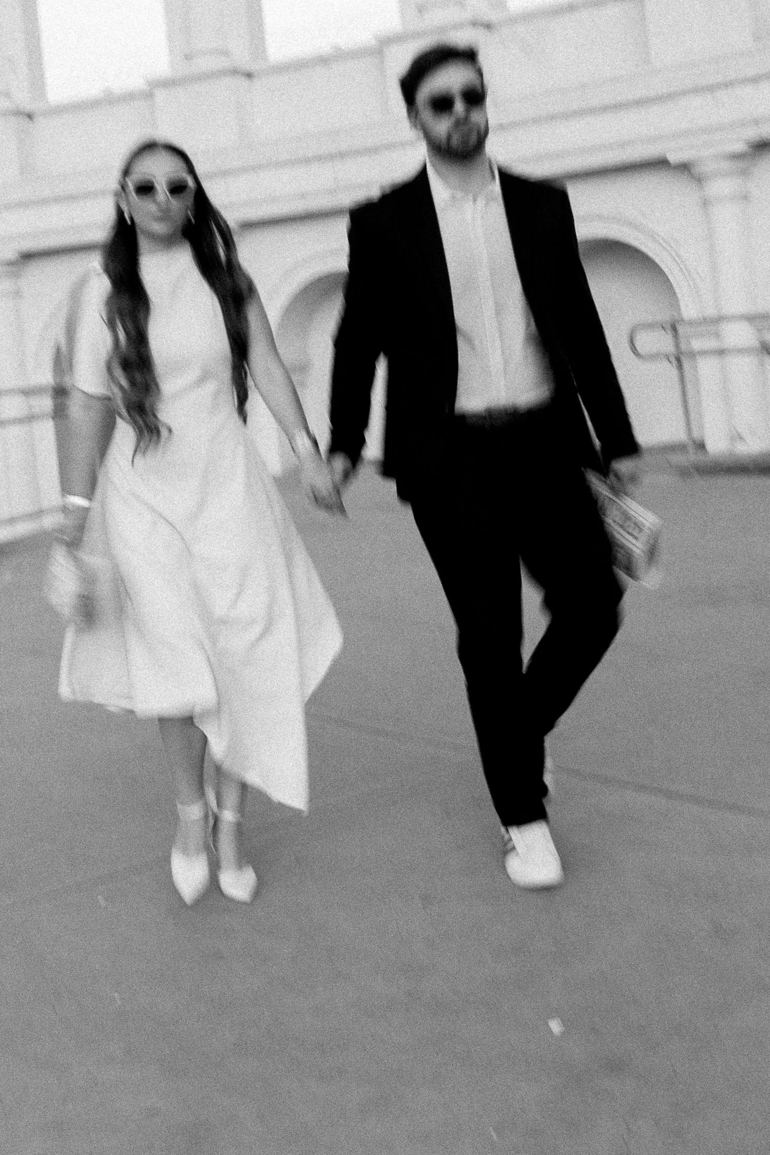 Black and white photo of a couple holding hands walking, woman in white dress and man in black suit wearing sunglasses.