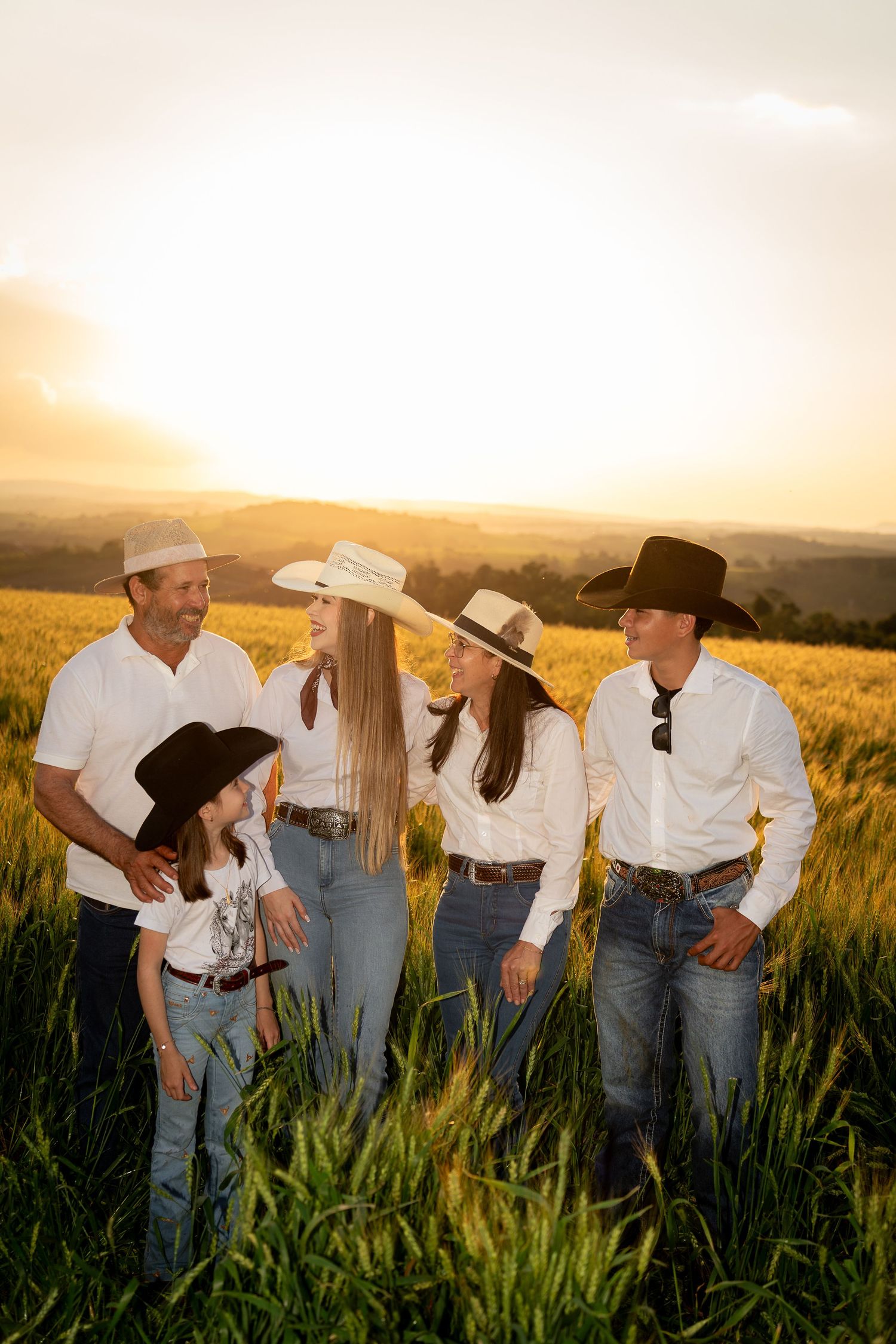 Family of five in white shirts and cowboy hats standing in a golden wheat field at sunset.