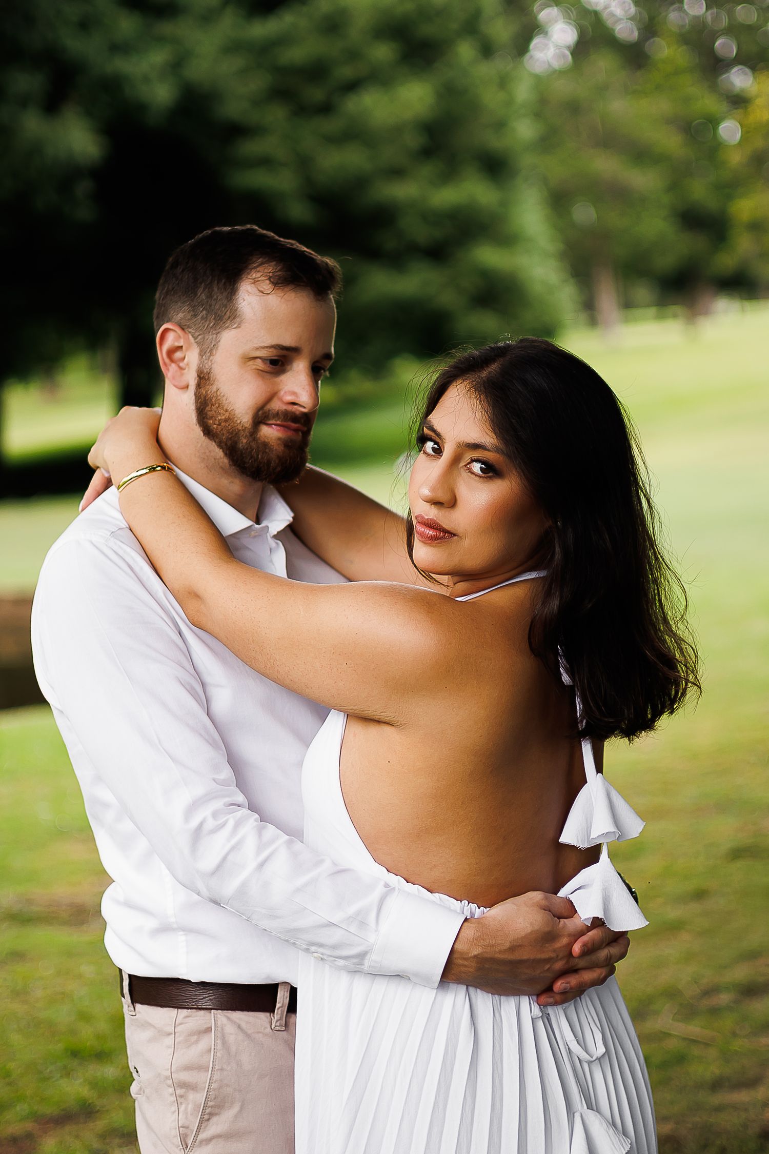 Couple in white outfits embracing outdoors, woman in backless dress looking at camera, lush green trees in background.