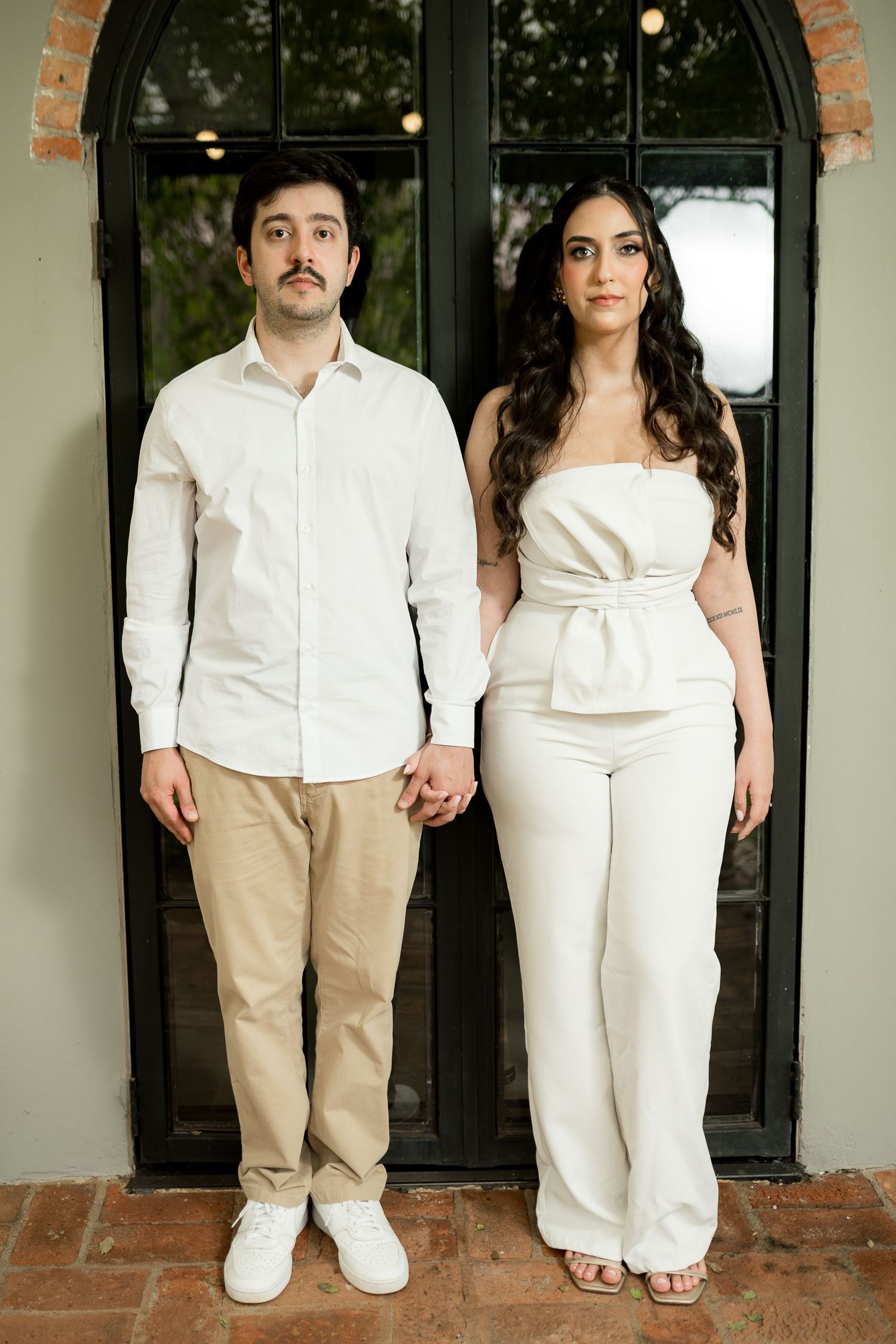 Couple holding hands in white outfits standing before a dark arched door, elegant engagement photo.