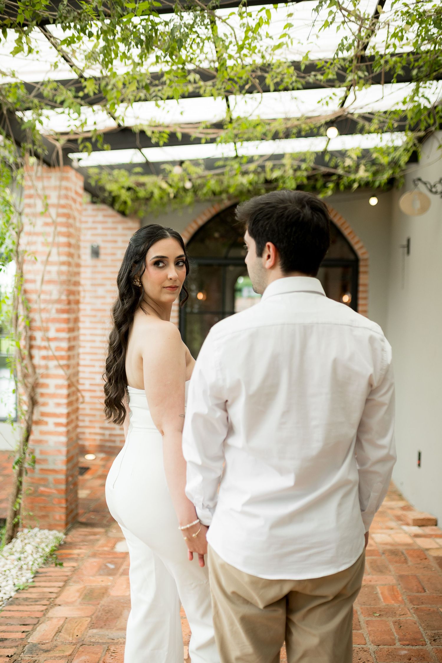 Bride in backless white gown holds groom's hand under vine-covered pergola at outdoor wedding venue.
