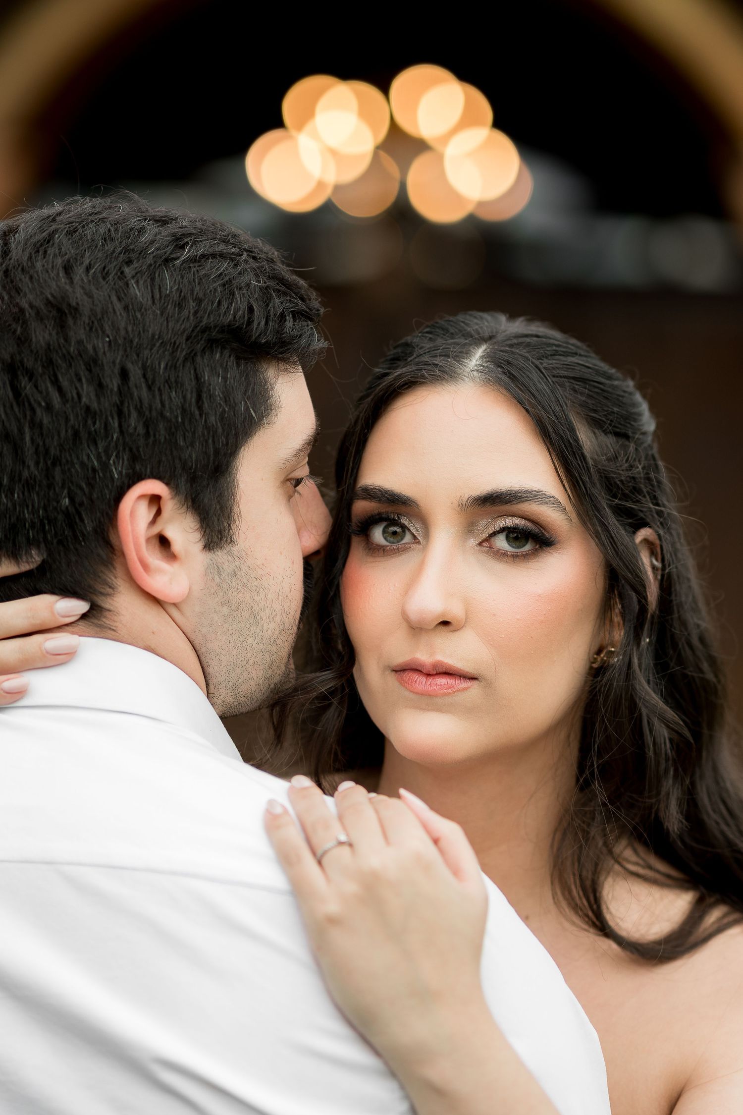A bride with dark hair and smoky eye makeup gazes forward as groom whispers in her ear during romantic wedding portrait.