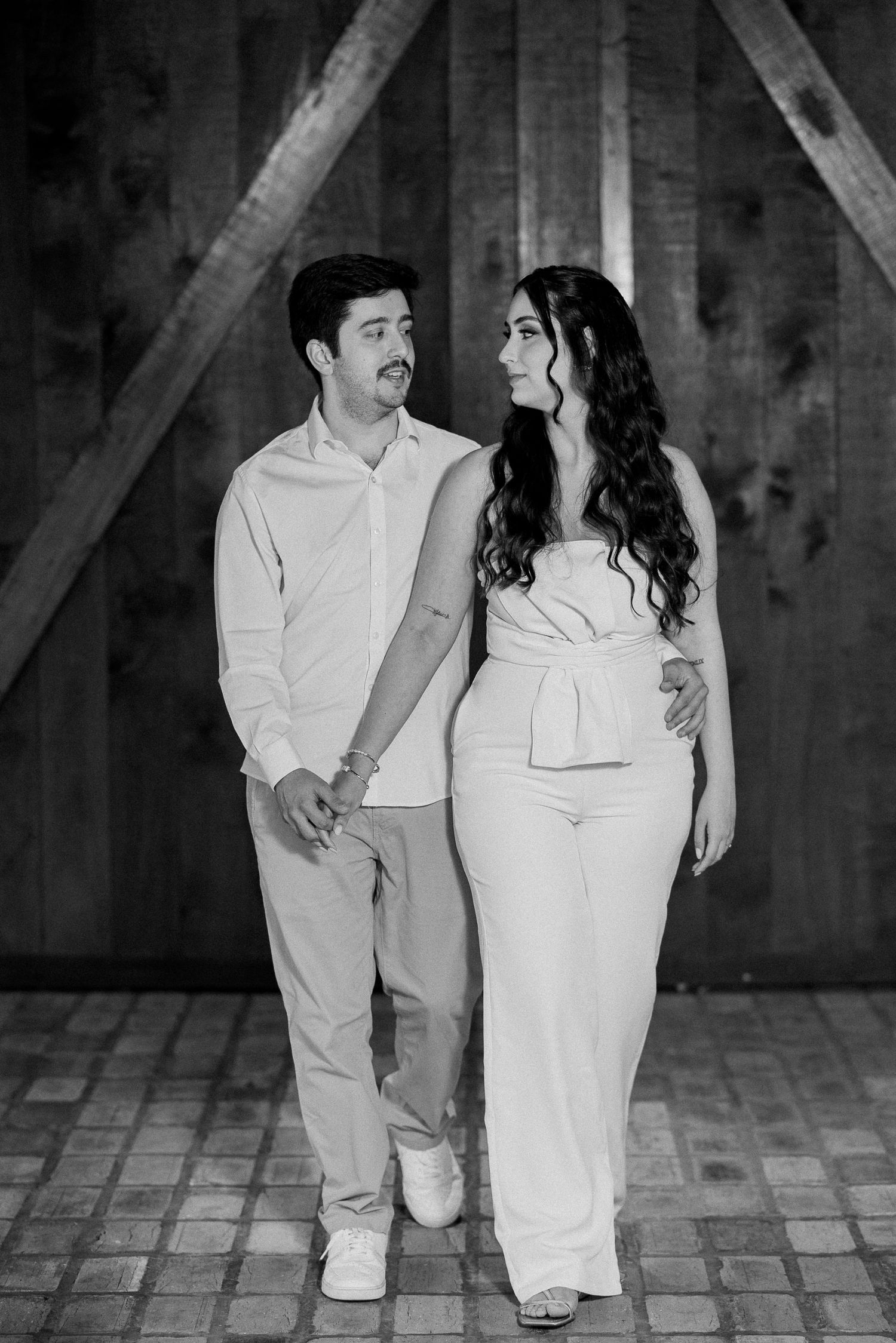 Black and white photo of a couple holding hands and gazing at each other in front of a rustic wooden barn wall.