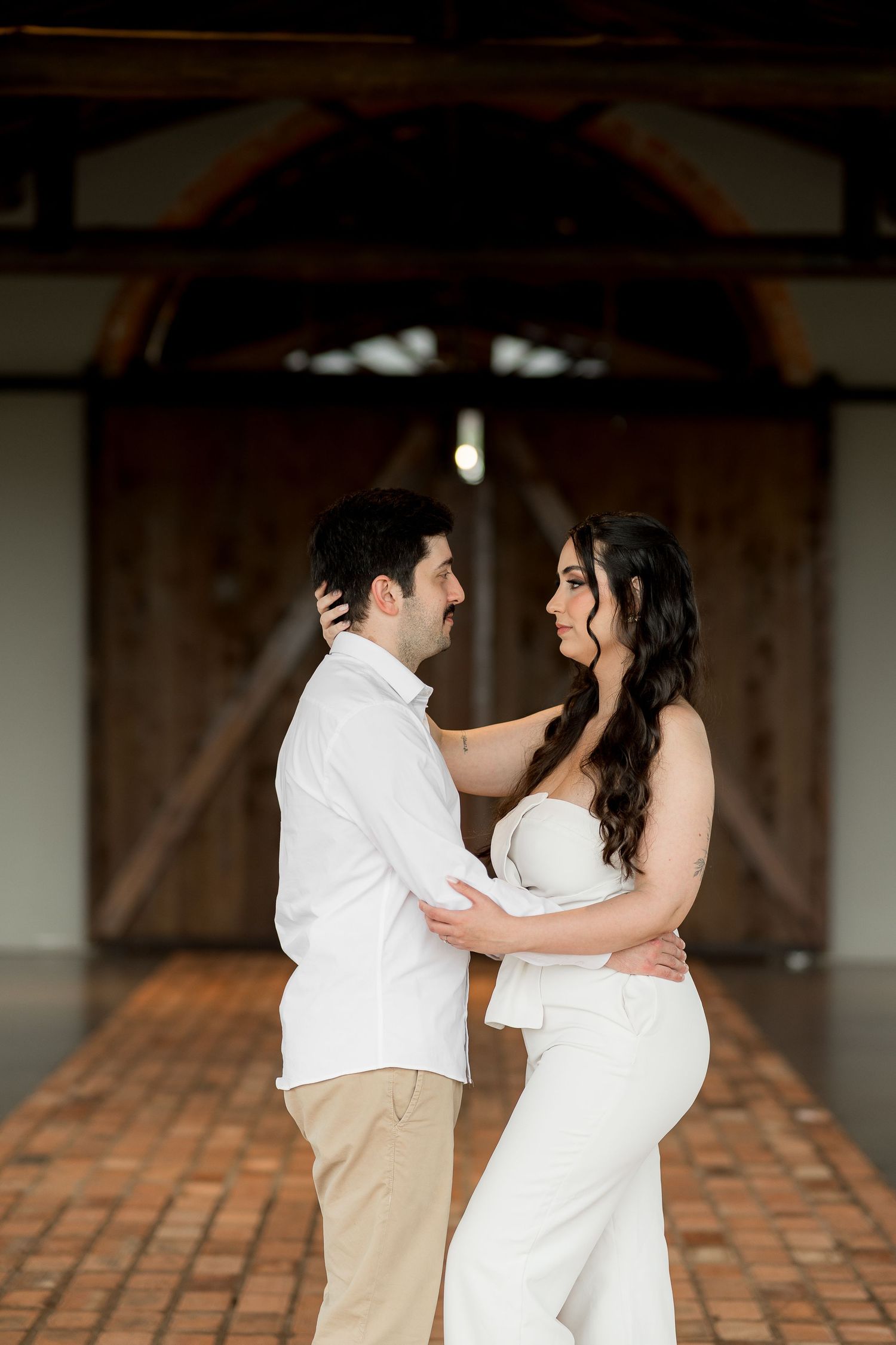 Couple in white attire embracing and smiling at each other inside a rustic barn with wooden doors.