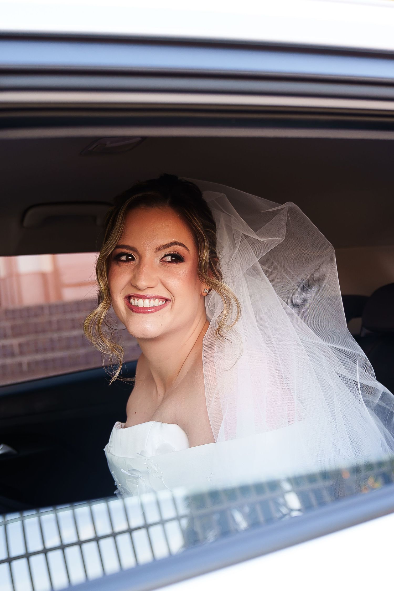 Smiling bride in white veil and gown sitting in a car window on her wedding day.